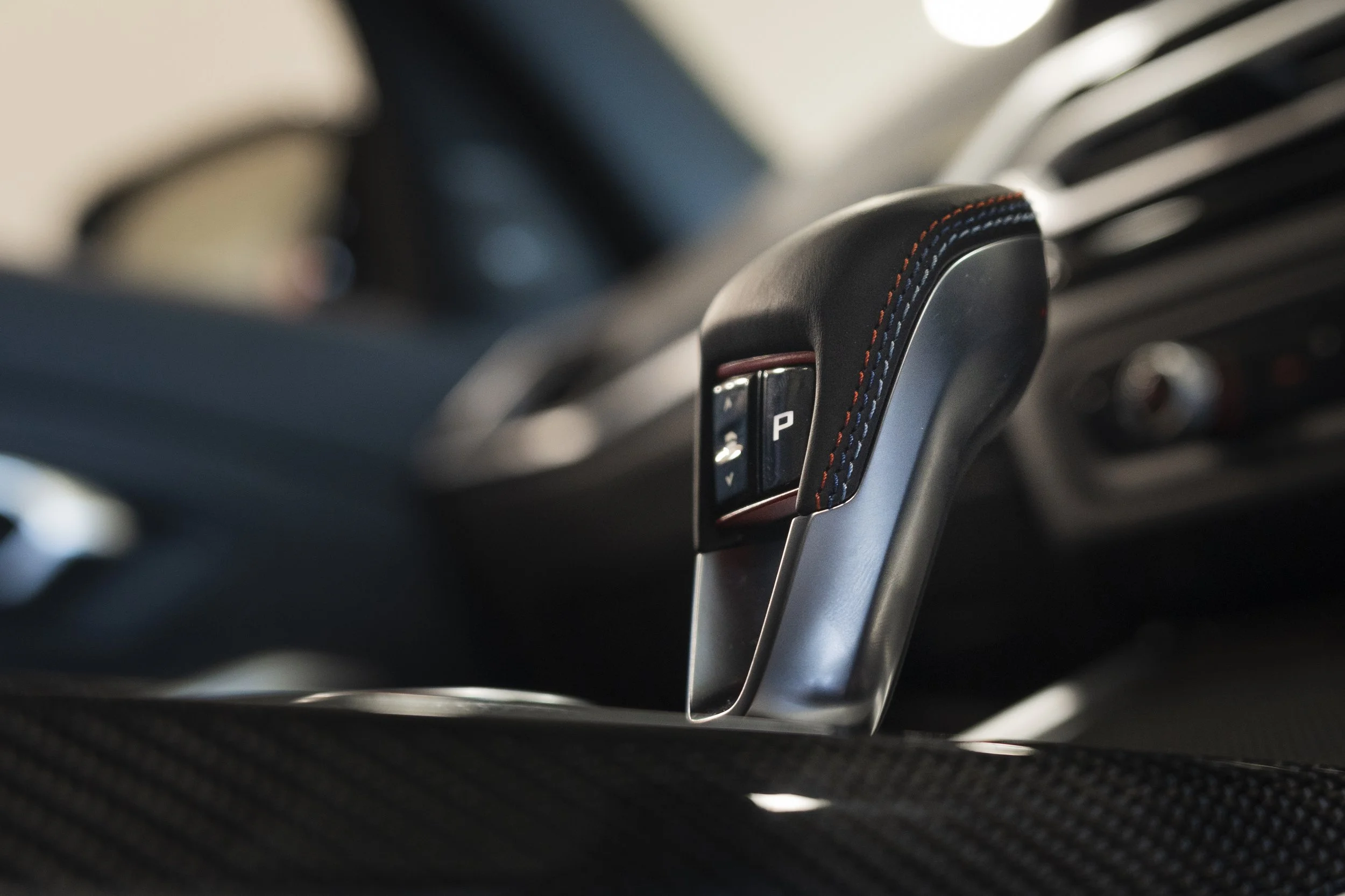 Close-up of a car gear shift lever with black leather and multi-colored stitching, marked with a 'P' for park, inside a vehicle.