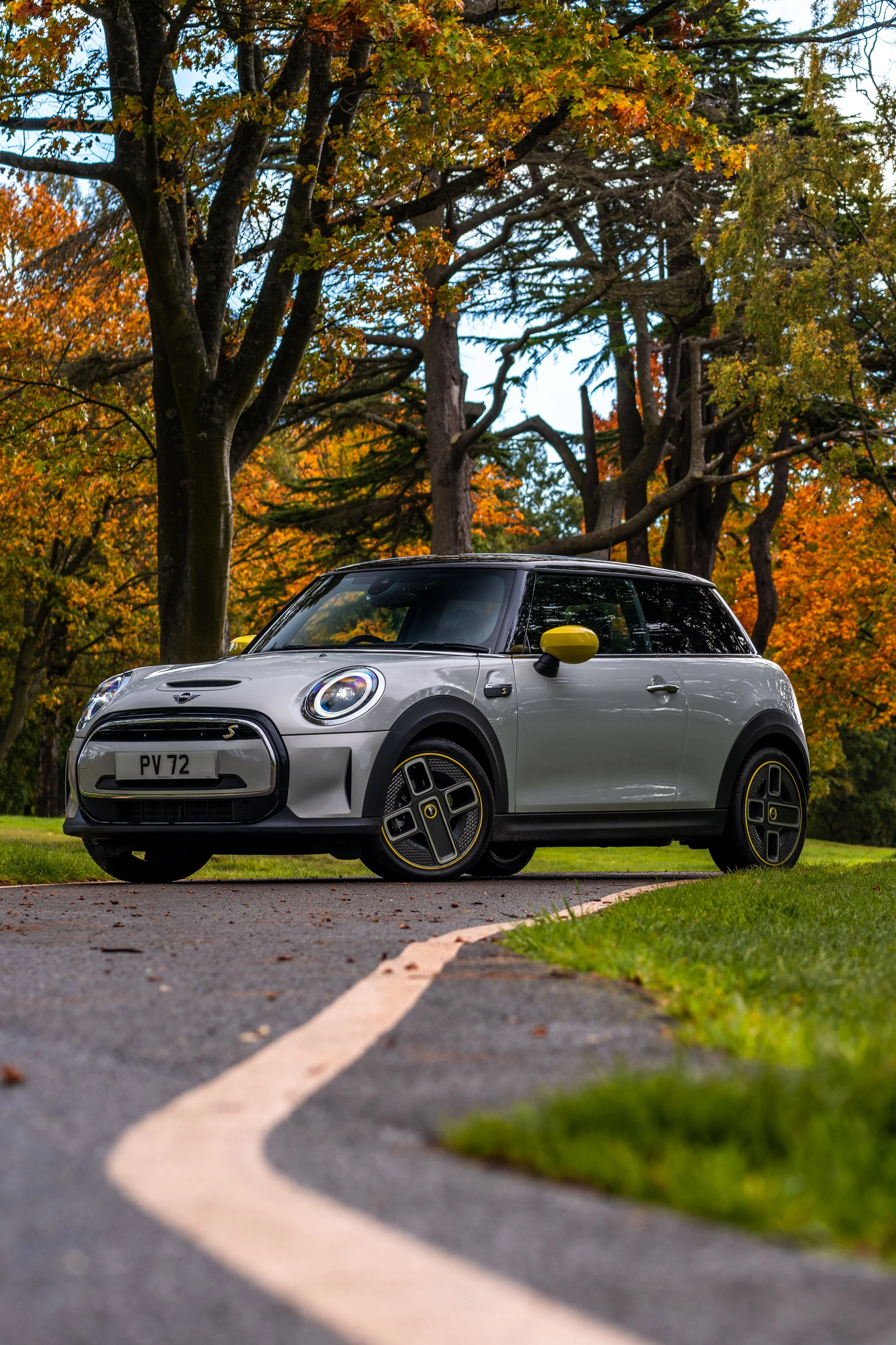 A silver Mini Cooper Electric parked on a paved driveway with a tree-lined background displaying fall foliage in orange and yellow colors.