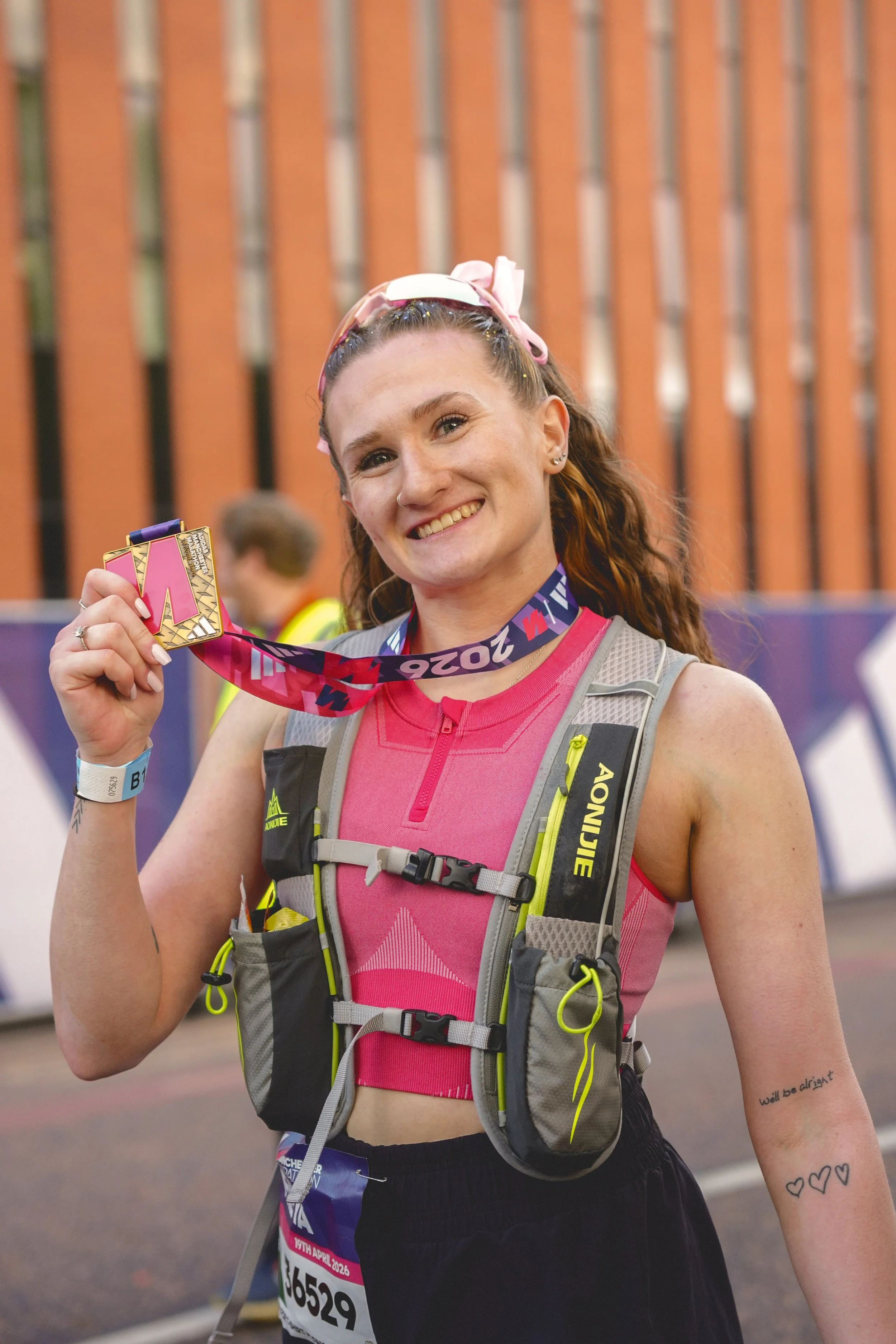 A smiling woman holding a medal at a race event, wearing running gear including a pink top, a hydration vest, and a race bib, with a tattoo on her arm reading "we'll be alright" and three hearts.