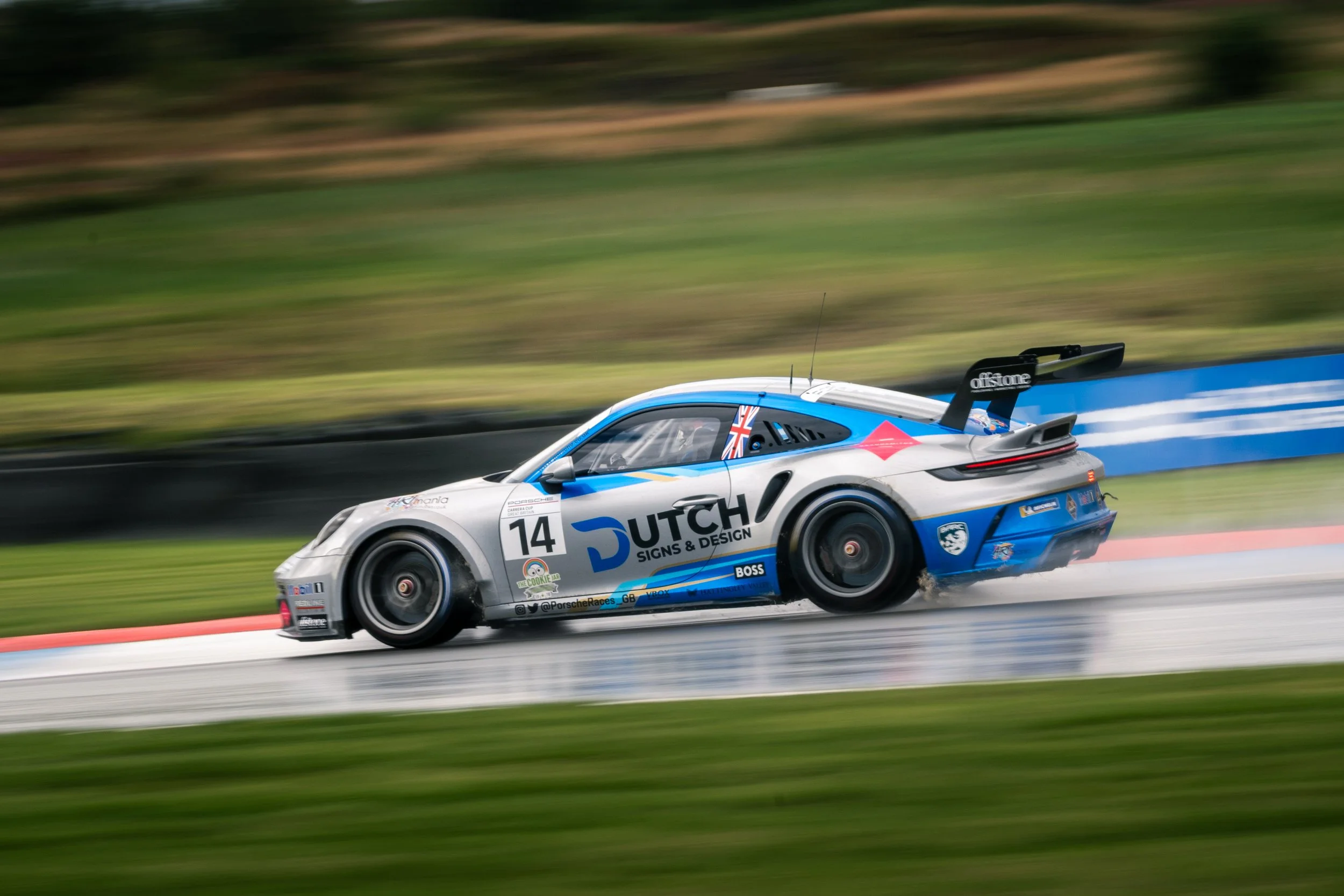 A racing car with the number 14, painted in blue, white, and gray, speeding on a wet race track with spray behind it, during a race. Knockhill racing circuit during porsche carrera cup.