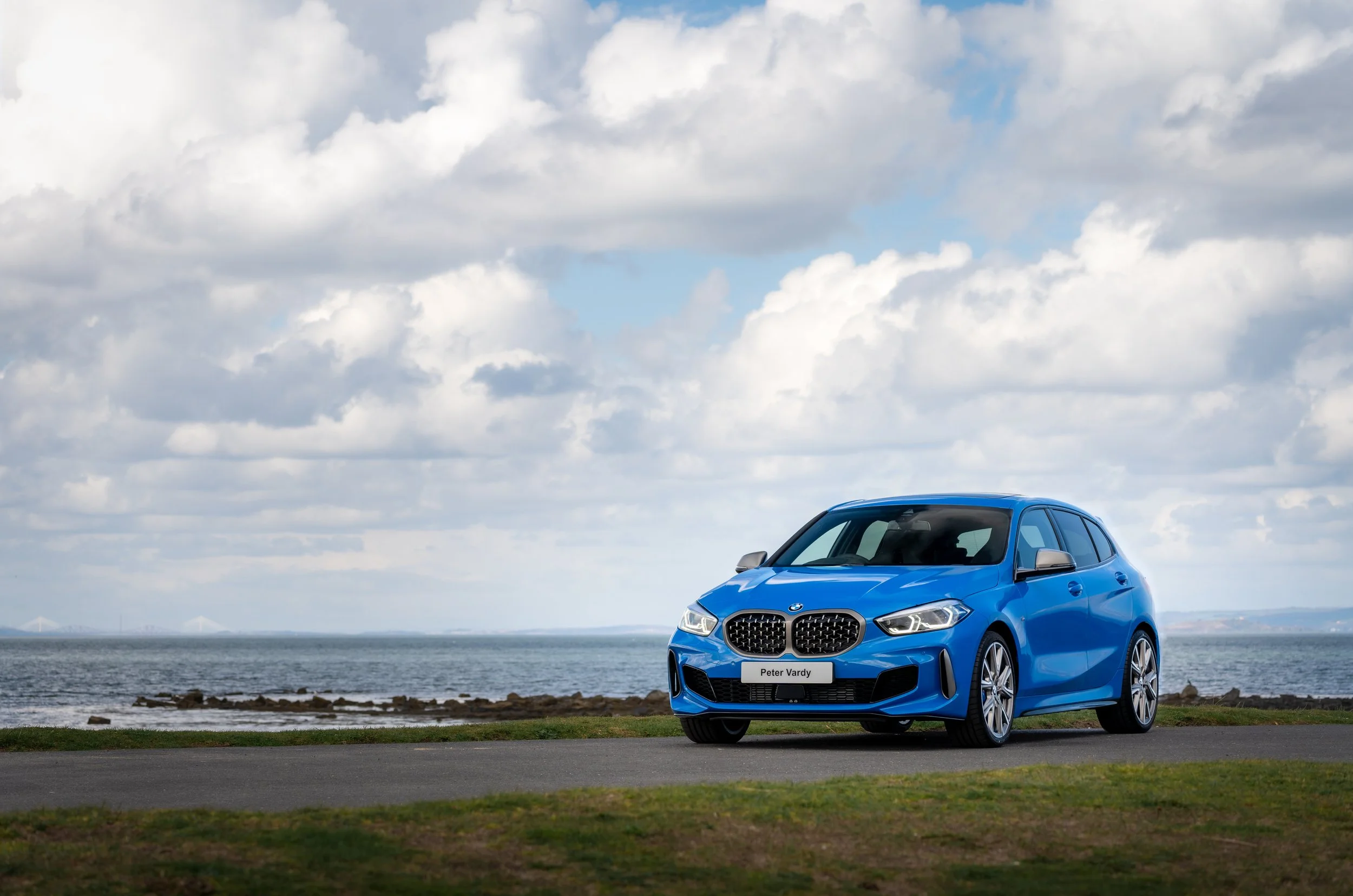 Blue BM1 Series car parked near a grassy area by the ocean under a cloudy sky.