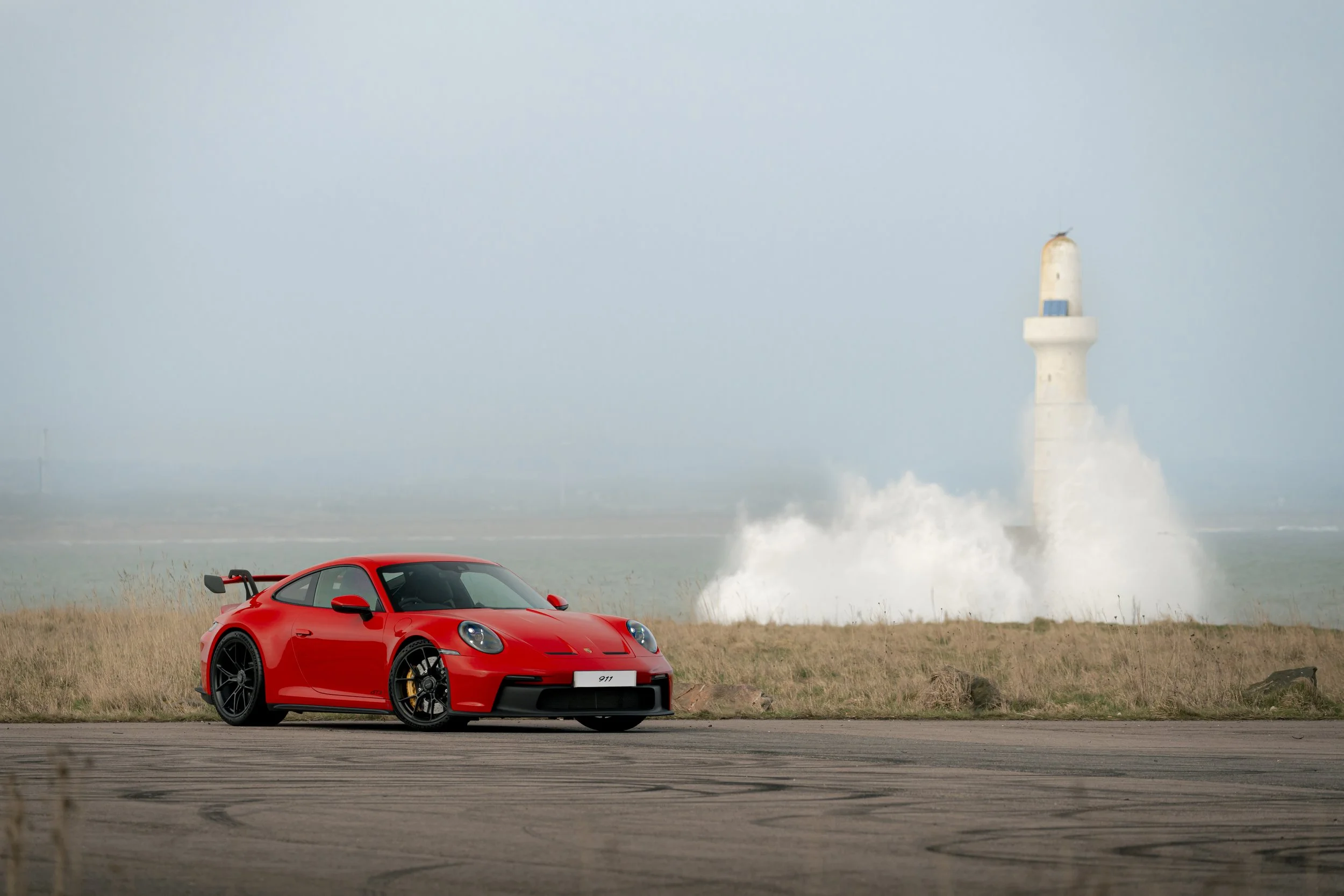 Red Porsche Gt3 car on a runway near a lighthouse with waves crashing at the base.