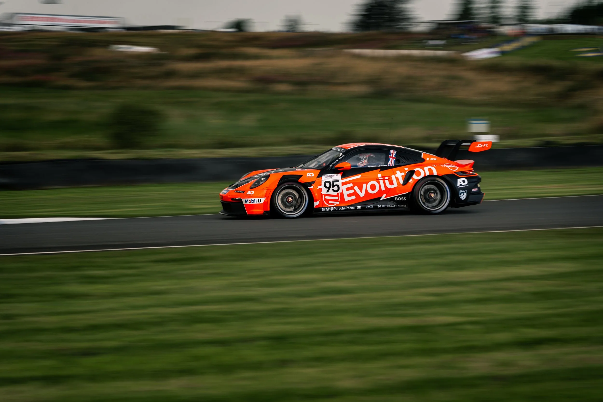 Race car with orange and black livery, number 95, moving on racetrack with blurred background. Porsche Carrera Cup at Knockhill