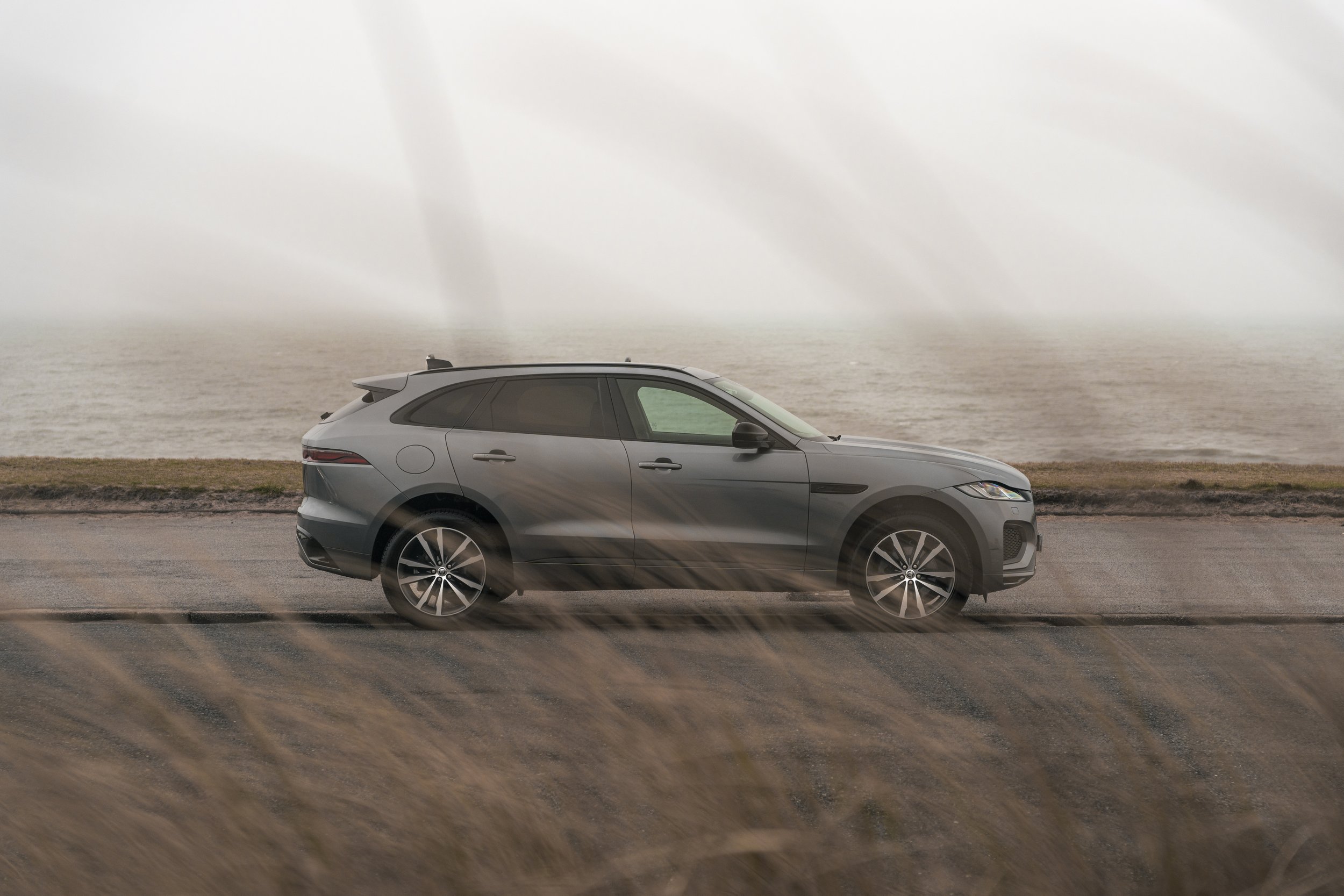A gray Jaguar F-pace SUV parked on a road by the ocean with grass in the foreground and a cloudy sky in the background.