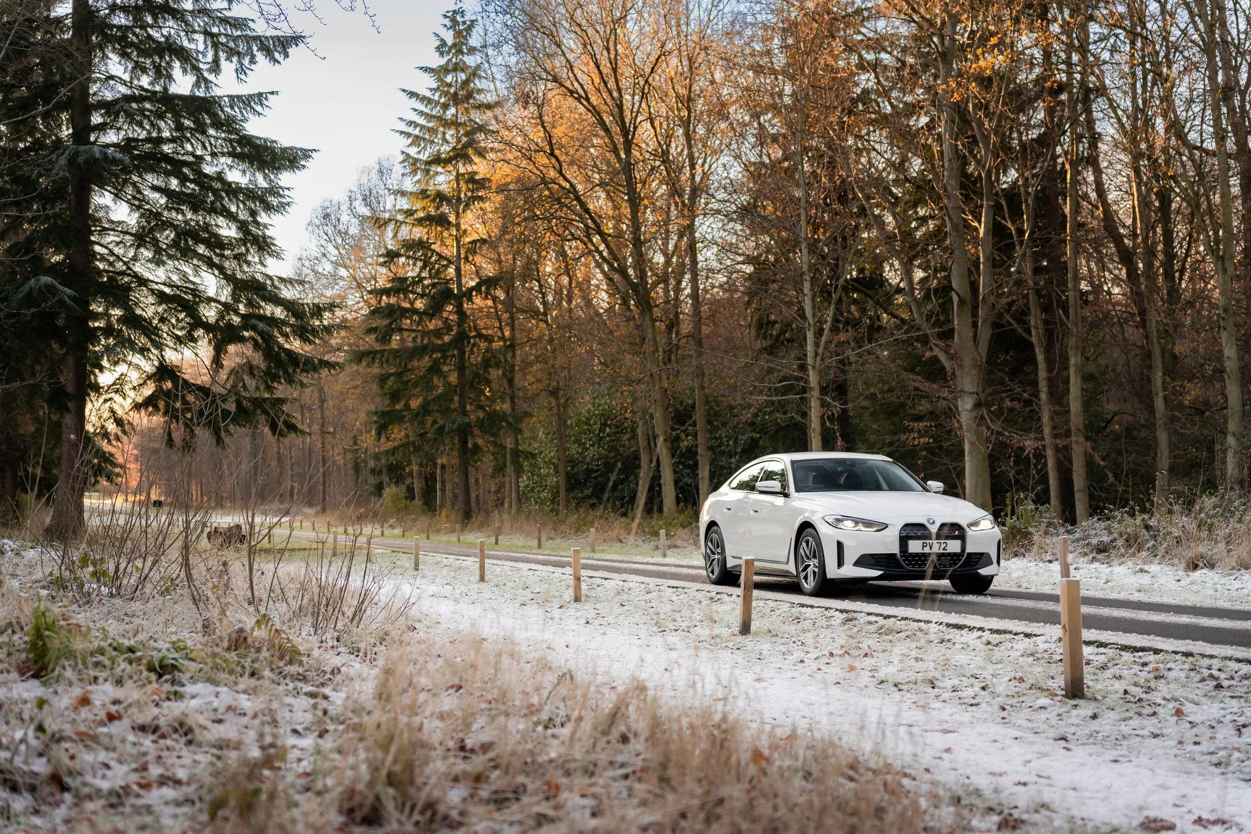 A BMW i4 car parked on a rural road surrounded by leafless trees and frosty grass, with a sunset sky in the background.