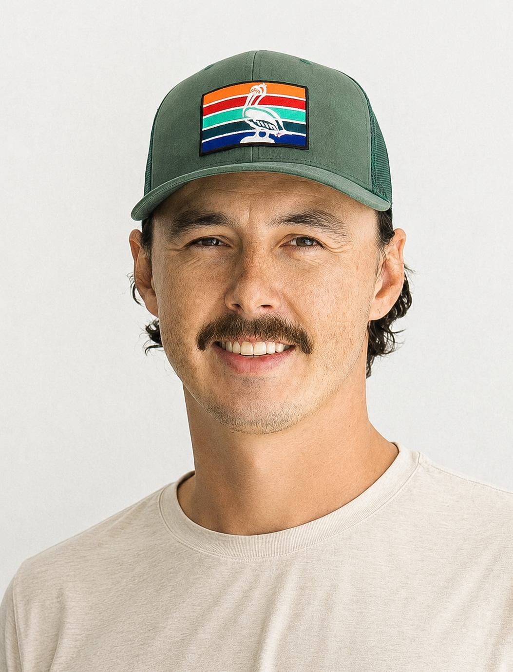 A smiling man wearing a beige T-shirt and a green cap with a rainbow-striped patch featuring a white bird illustration.