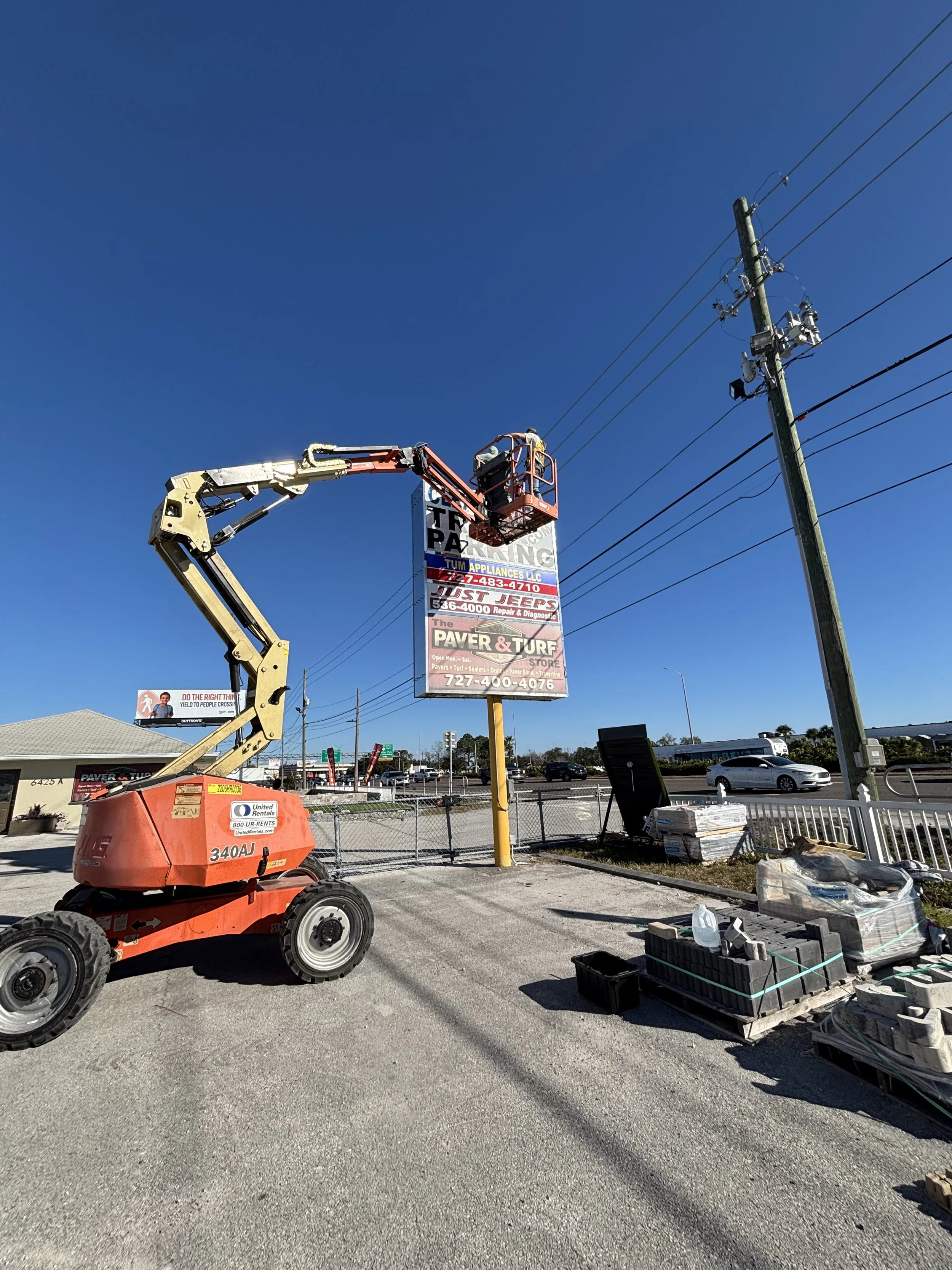 Construction equipment with a lift basket working near power lines, a utility pole, a sign for paving and turf services, and a parking lot with cars in the background under a clear blue sky.