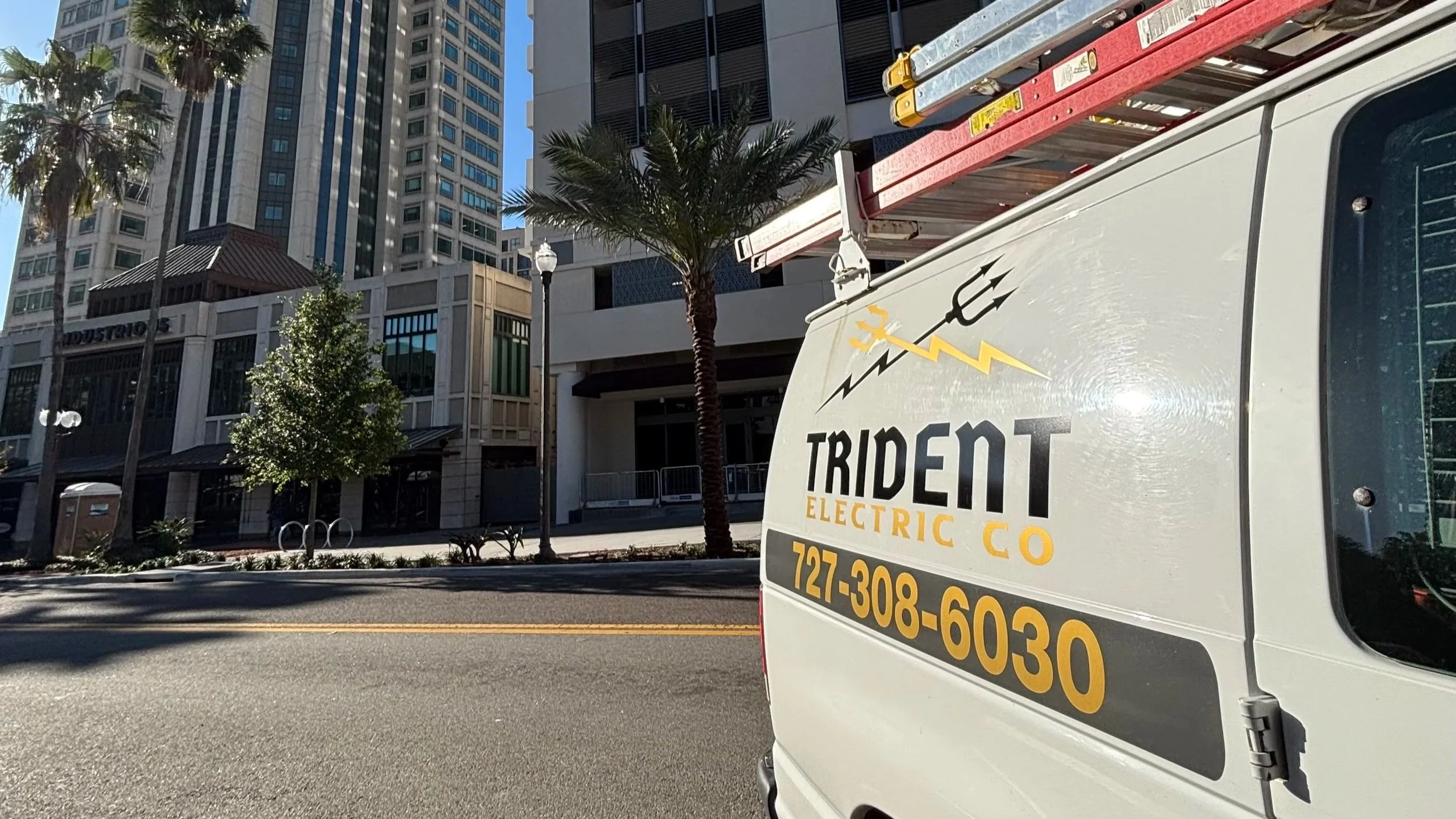 City street scene with high-rise buildings, palm trees, and a utility truck with company branding and contact information parked near the sidewalk.