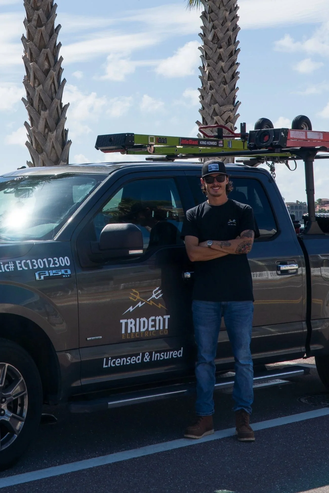 A man in sunglasses and a cap standing with arms crossed beside a gray utility truck with a ladder on top, in a parking lot with palm trees and a partly cloudy sky in the background.