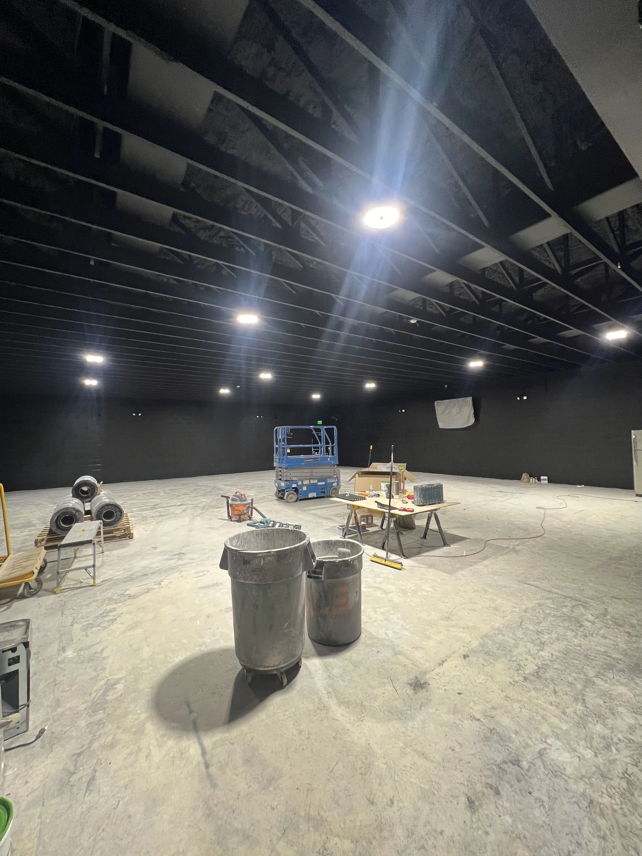A large room under construction or renovation with overhead lighting and construction tools and materials scattered on the floor, including trash bins, a lift, and work tables.