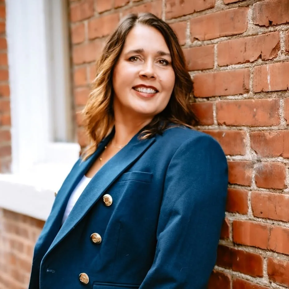 Professional headshot of a woman with shoulder-length brown hair, wearing a black blazer over a blue top, smiling at the camera, with a blurred indoor background.