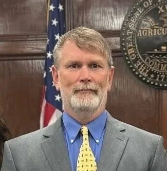 A smiling man in a dark suit, white shirt, and a navy blue striped tie, standing in front of an American flag.