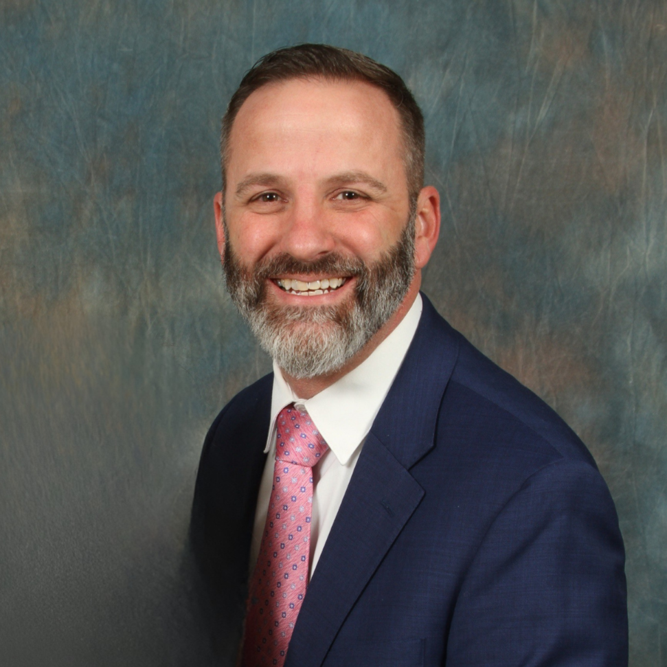 A smiling man with a beard, wearing a navy suit, white shirt, and pink tie, in front of a textured background.