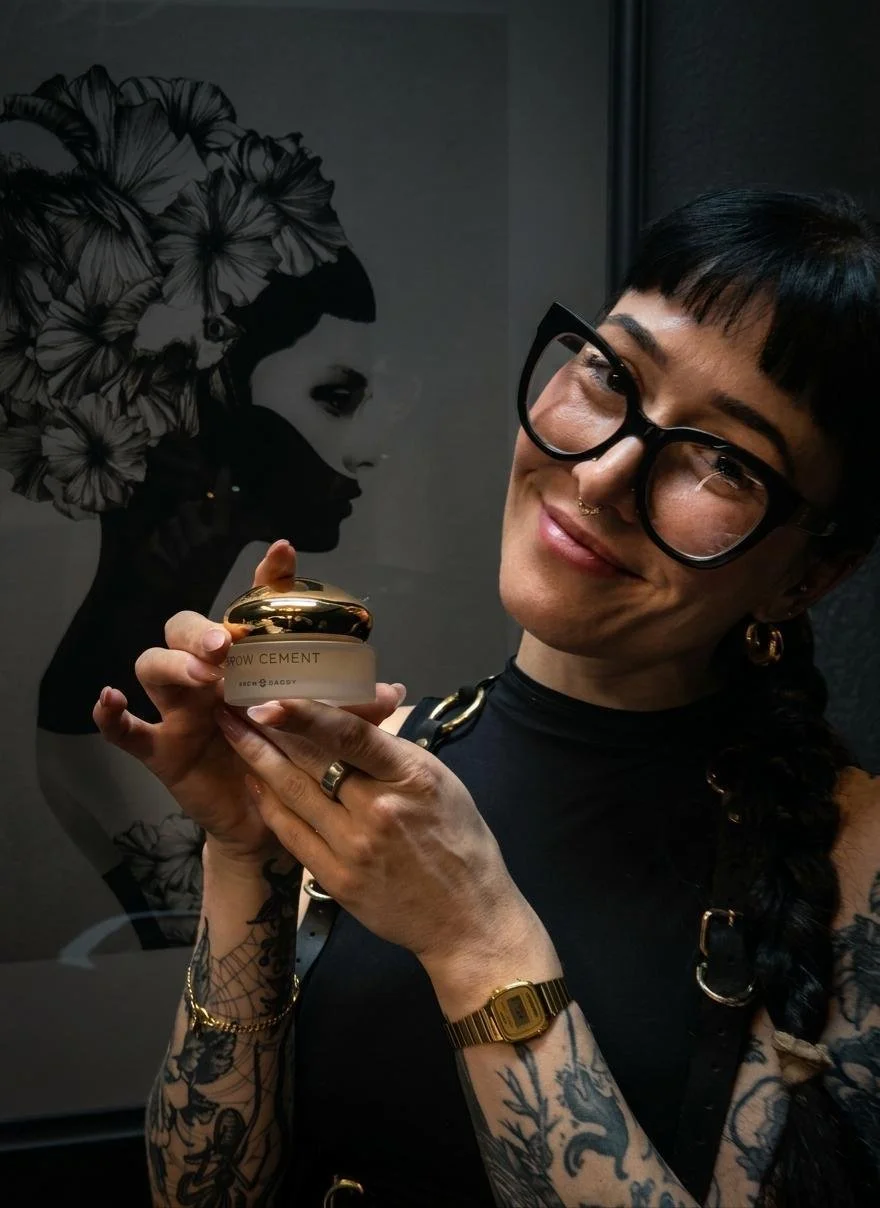 A woman with short black hair, glasses, and tattoos is holding a jar of 'Glow Cement' skincare product, smiling at the camera. Behind her is artwork of a woman with flowers in her hair.