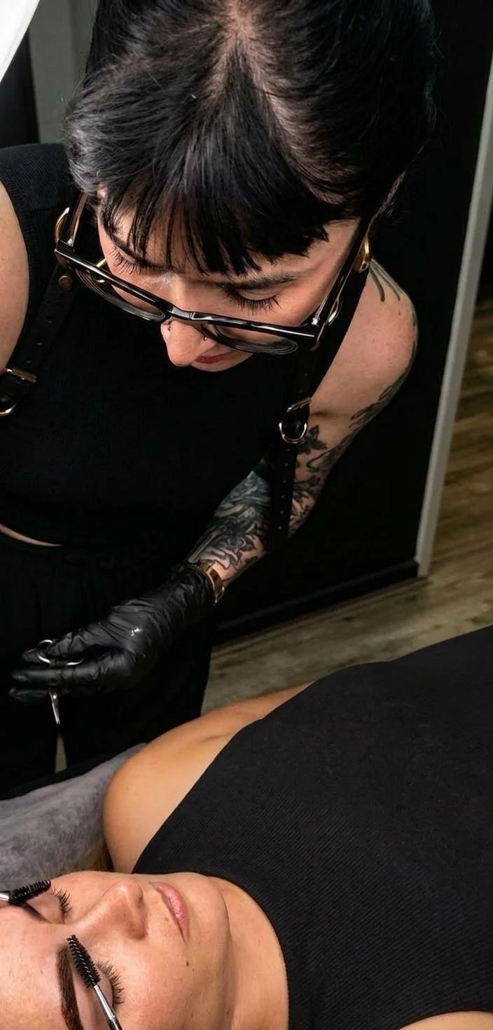 A woman lying on her back with eyelash extensions applied by a tattooed artist wearing black gloves.