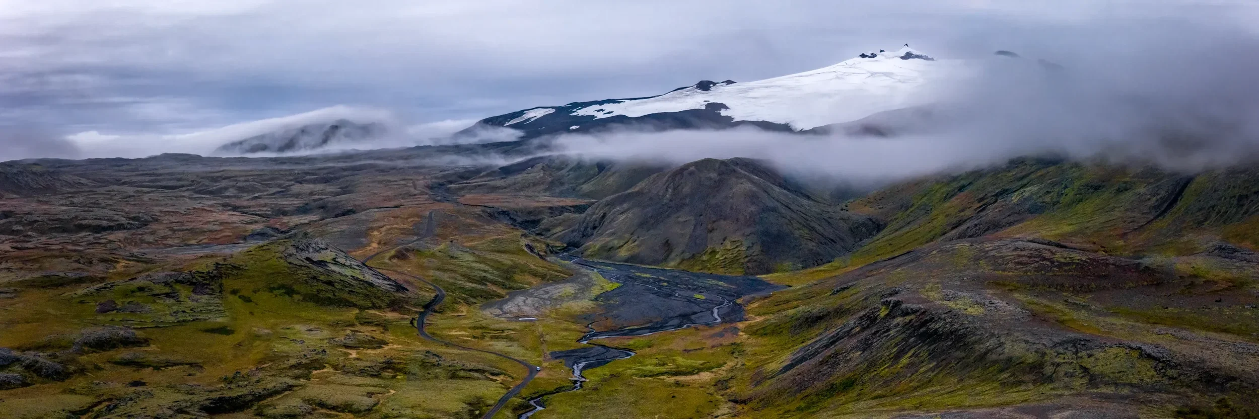 The Snaefellsjokull valley with Snaefellsjokull glacier in the clouds. A scenic landscape featuring rolling green hills, winding roads, and streams in the foreground with snow-capped mountains and clouds in the background.