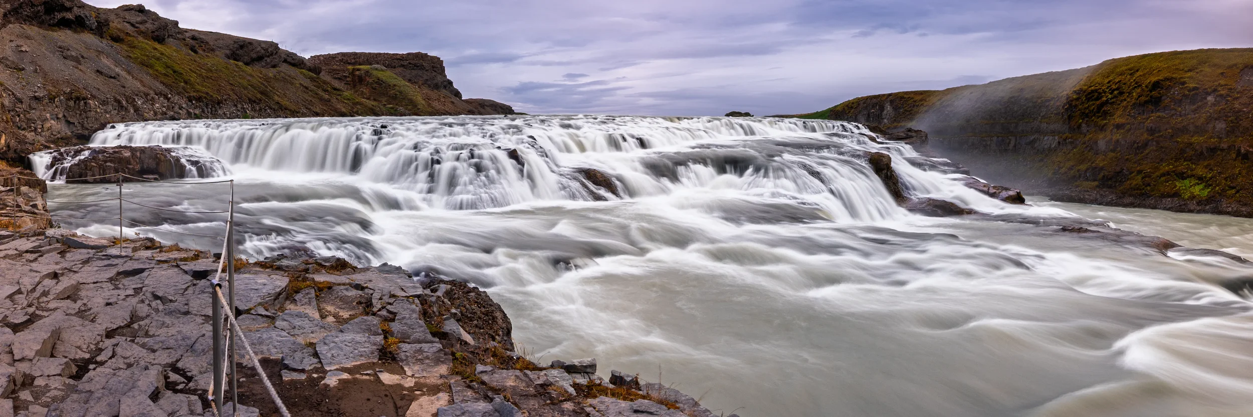 Gullfoss in Iceland flows over the upper waterfall, with silky long exposure water flowing over rocks, under a cloudy stormy sky, surrounded by autumn hills.