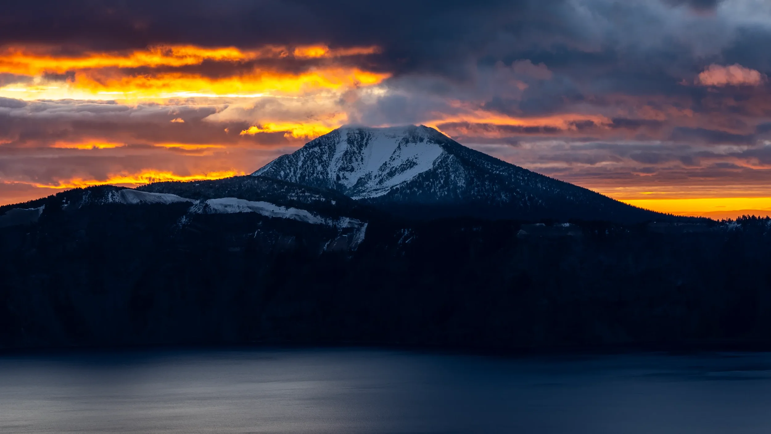 Mt. Scott at Crater Lake with a dusting of snow under a dramatic orange and purple sunset with dark clouds and a calm body of water in the foreground.