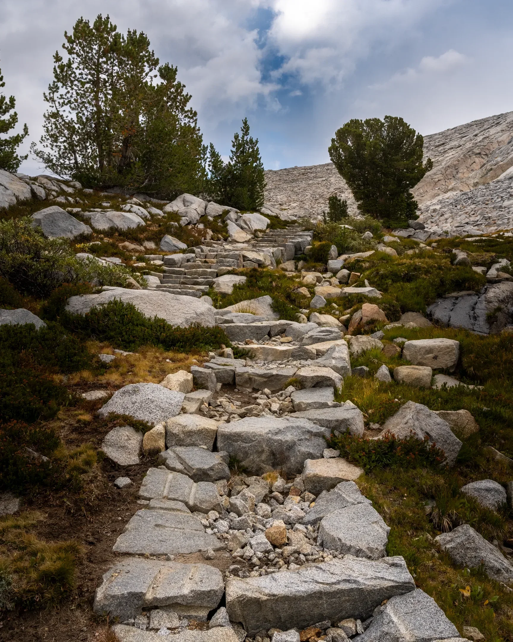 Stone steps ascend along the JMT approaching Donahue Pass while hiking southbound (SOBO), with a rocky hillside with pine trees and a cloudy sky.