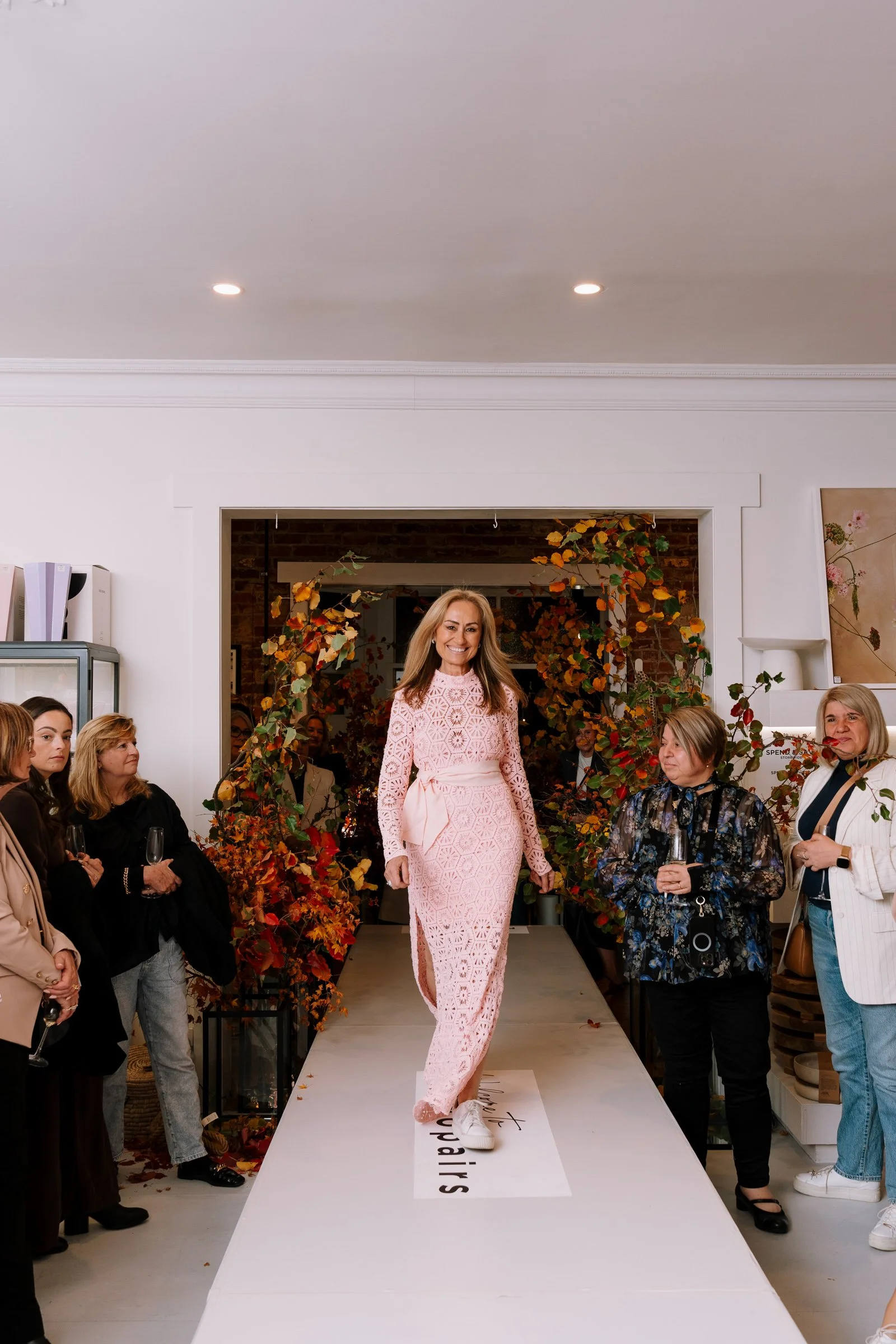 A woman in a pink lace dress walking on a runway at a fashion event, surrounded by seated and standing attendees in a decorated indoor space.