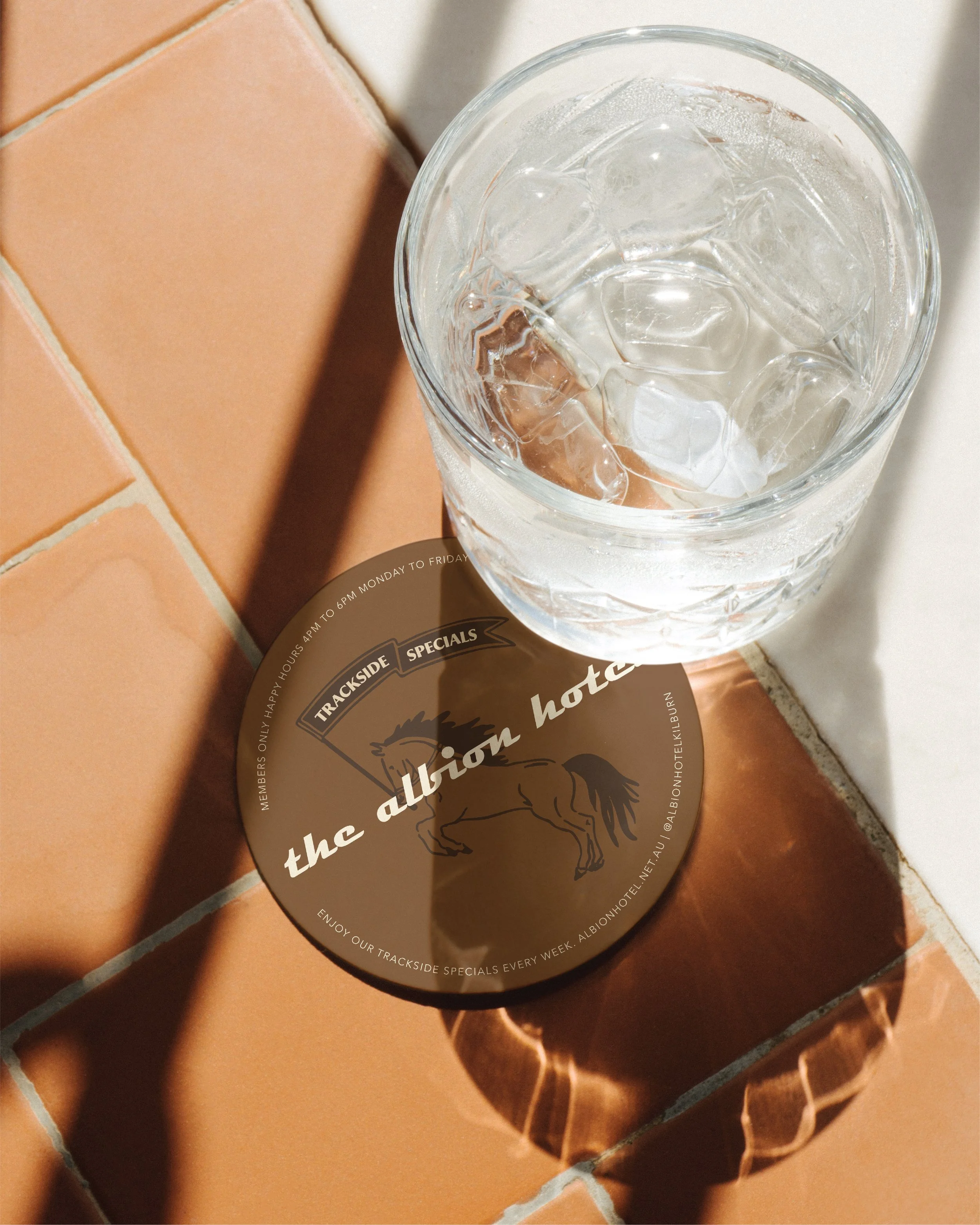 A glass of ice water on a coaster with a horse illustration and text for The Albion Hotel, placed on a tiled surface with shadows.