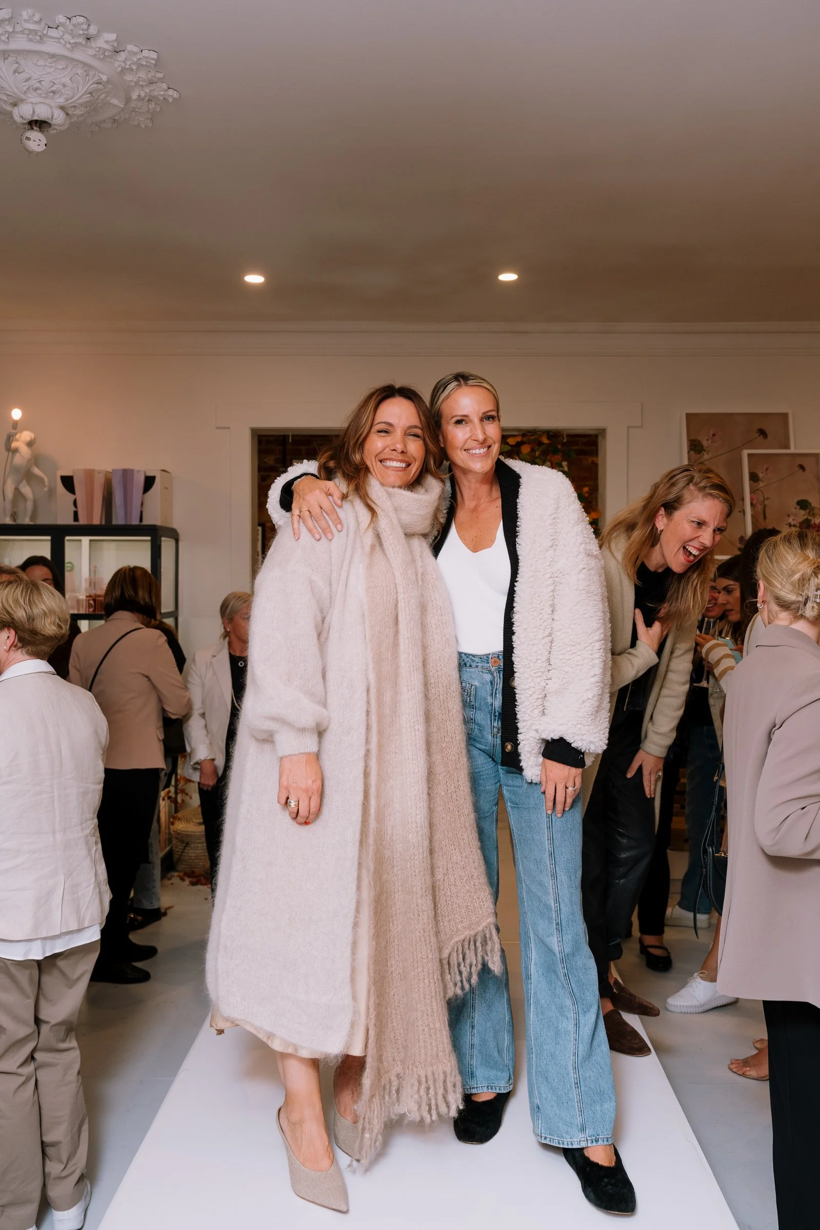 Two women standing close together, smiling, with a group of people socializing in the background, in an art gallery or event space.