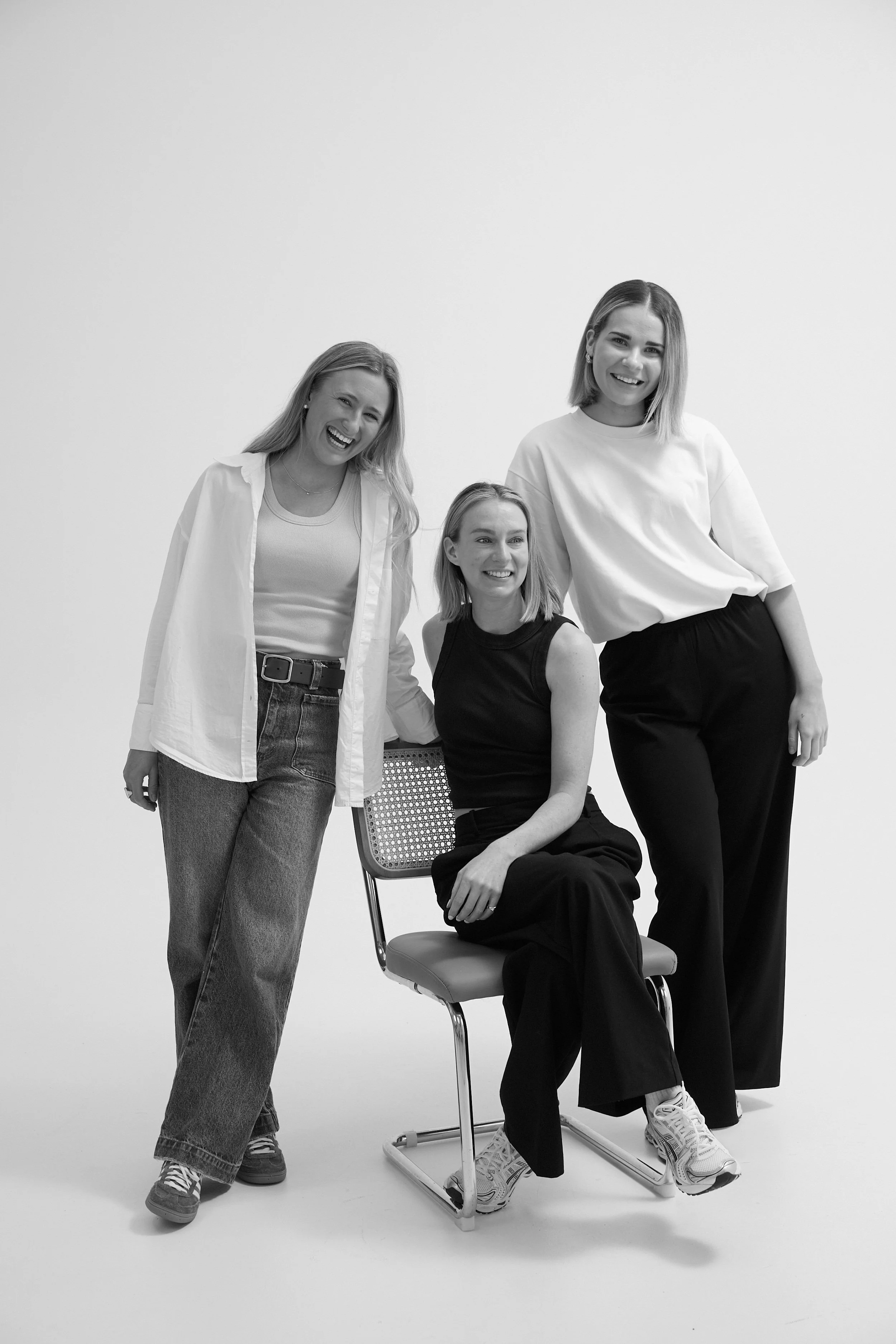 Black and white photo of three women smiling, with one sitting on a chair and two standing beside her against a plain background.