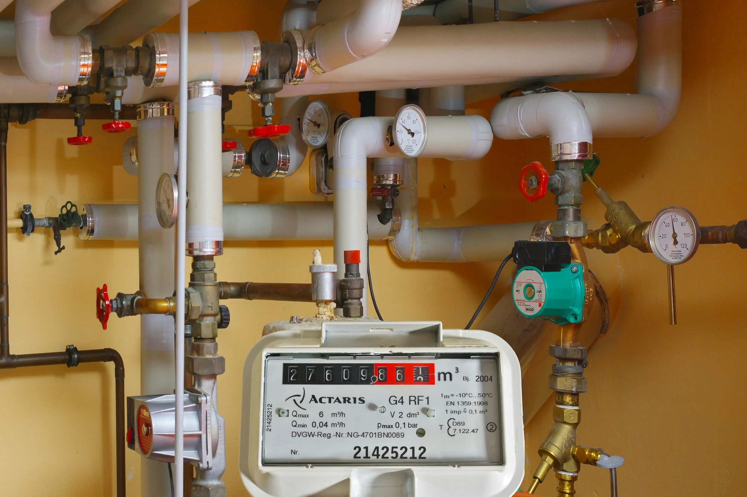 A water meter with pipes, valves, pressure gauges, and a pump in a plumbing system against a yellow wall.