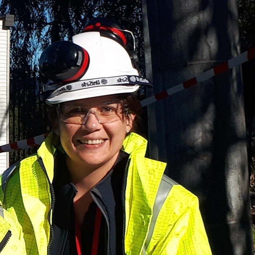 A smiling woman wearing a yellow safety jacket, clear glasses, and a white safety helmet with a headlamp, outdoors near steel pipes and trees.