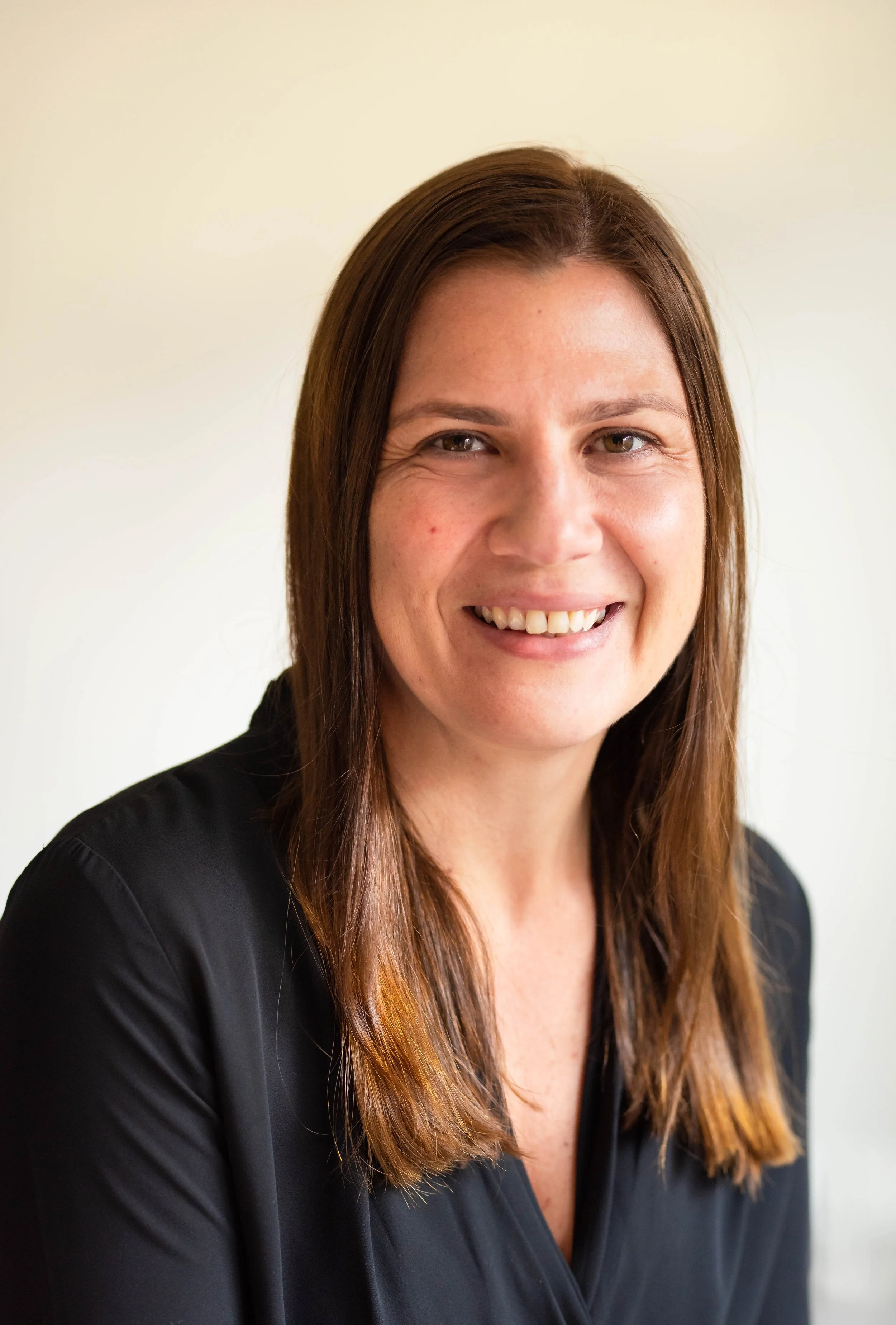 A smiling woman with shoulder-length brown hair wearing a black top, standing against a plain light background.