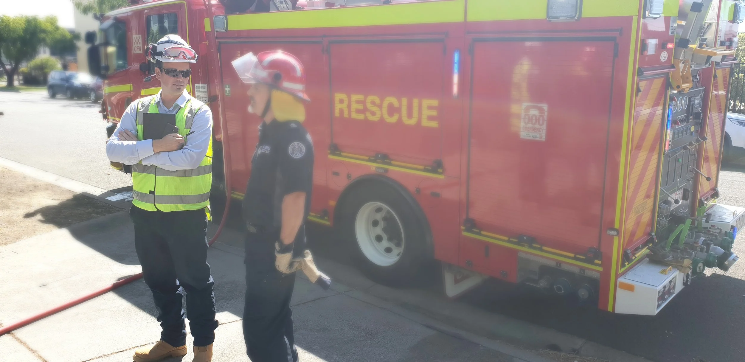 Two firefighters, one man and one woman, standing in front of a red rescue fire truck on a city street. The man wears a white shirt, reflective yellow safety vest, sunglasses, a helmet, and tan boots, holding a clipboard. The woman, dressed in a fire department uniform with a helmet, talks to him. The fire truck has the word 'RESCUE' written on the side and various firefighting equipment.