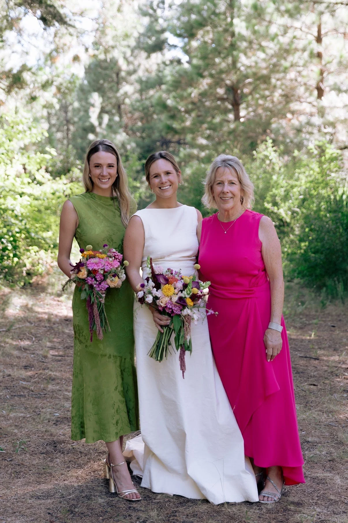 Three women standing outdoors in a wooded area, dressed in formal attire, holding bouquets of flowers, smiling at the camera.