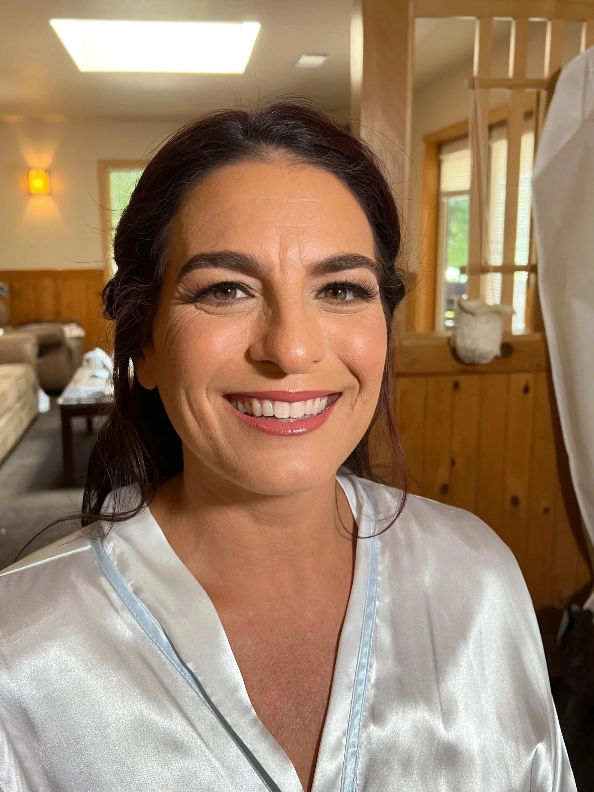 Close-up of a smiling woman with dark brown hair, wearing a white satin robe, indoors in a room with wooden paneling, a sofa, booths, and windows with blinds.