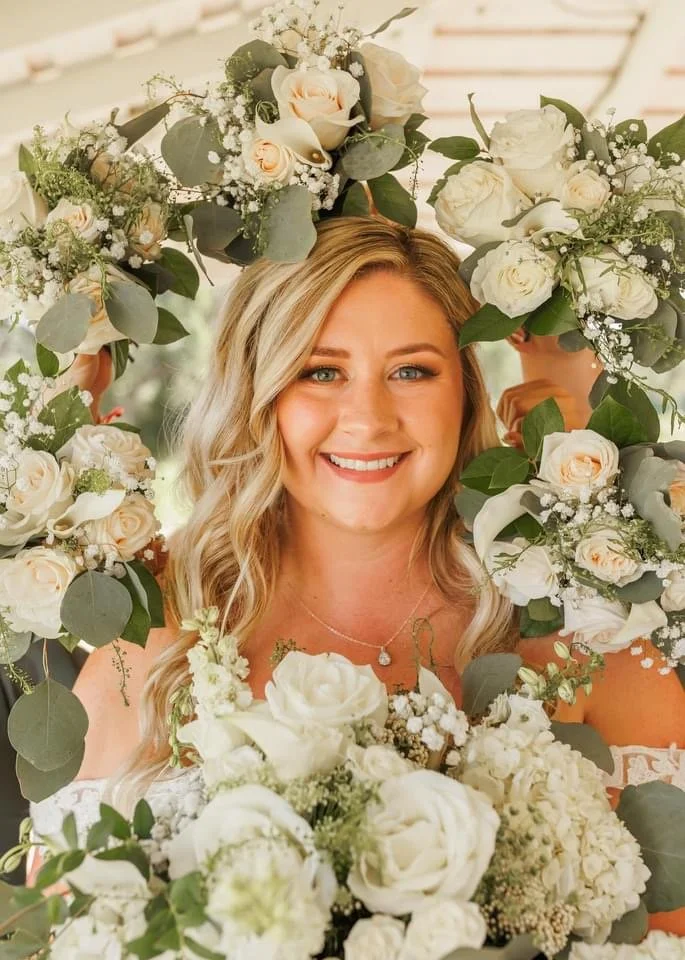 A smiling woman with blonde, wavy hair framed by a large floral wreath of white roses, baby's breath, and eucalyptus leaves, holding a bouquet of similar flowers.