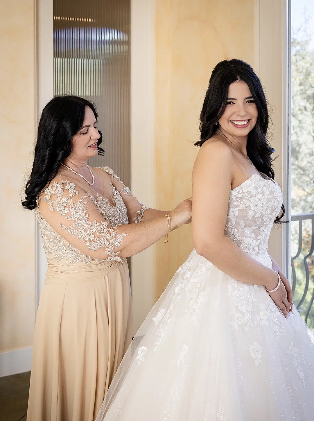 A woman in a white wedding dress smiling as another woman, likely her mother, helps her with her dress. The mother is wearing a beige dress with lace sleeves and a pearl necklace, standing in a room with yellow walls and a glass door.