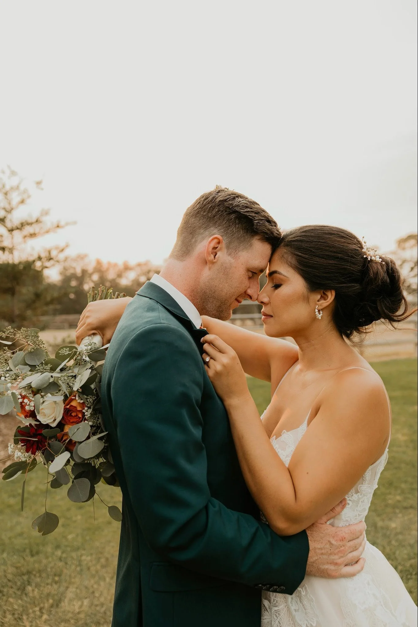 A bride and groom embrace closely outdoors during sunset, touching foreheads with eyes closed, holding a bouquet of flowers behind the groom.
