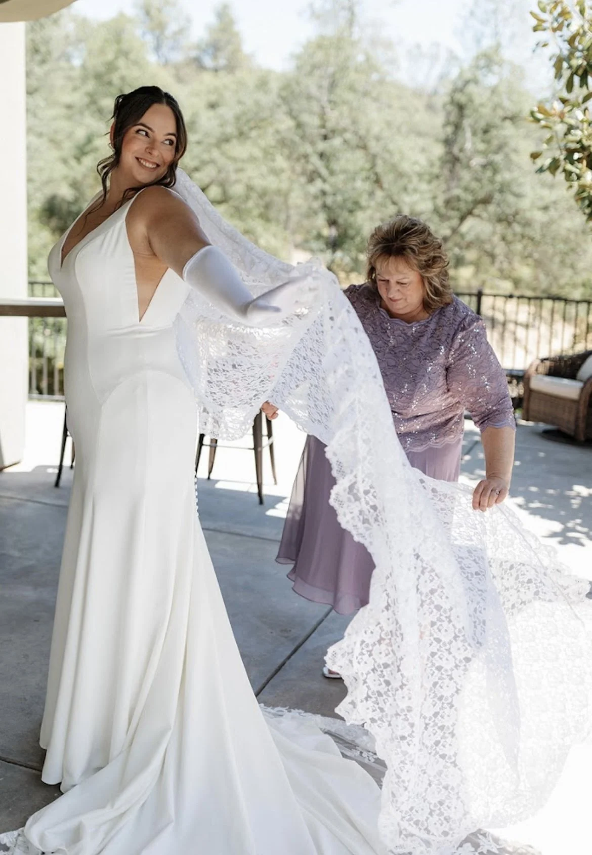 A bride with a joyful expression in a white wedding dress is being helped by an older woman to adjust her lace veil on an outdoor patio.