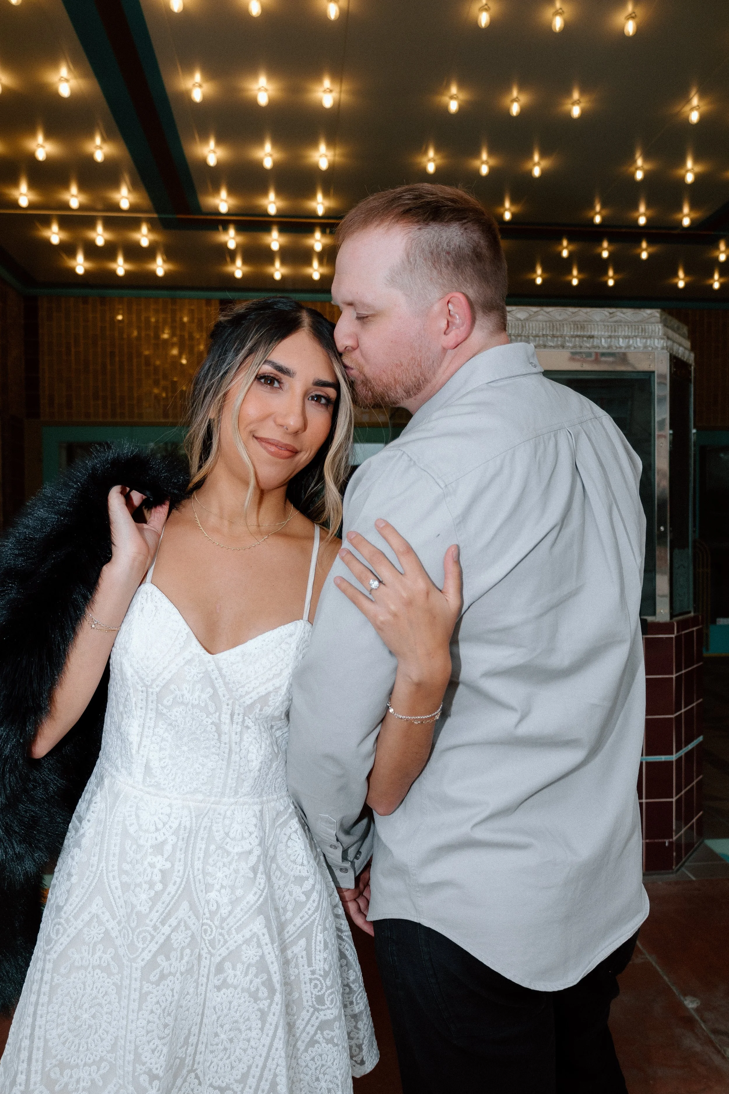 A couple stands close together in an indoor setting with string lights overhead. The woman is wearing a white lace dress, and the man is wearing a light-colored shirt. The woman has her hand on the man's shoulder, showing a wedding ring. The man is k