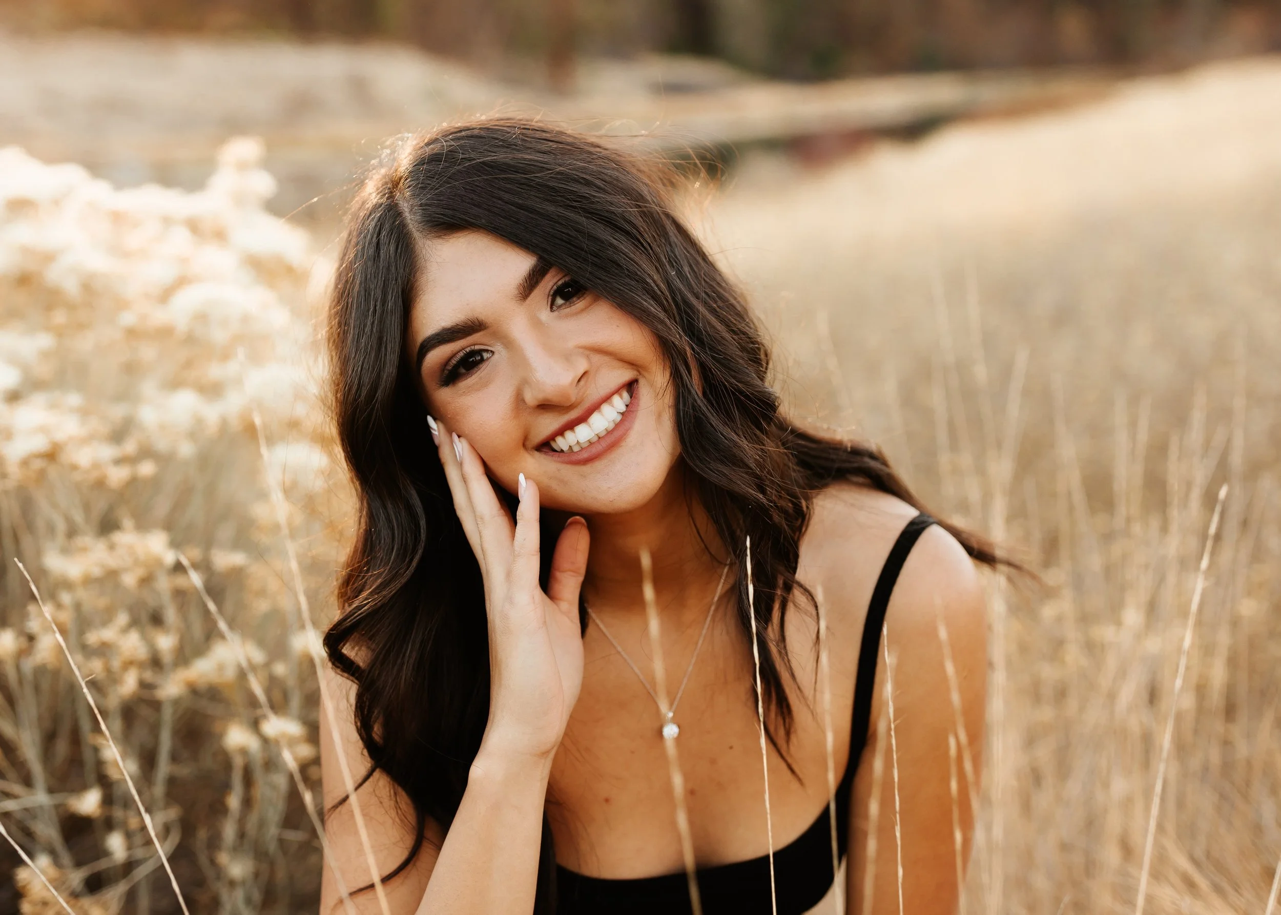 A young woman with dark, wavy hair smiling in a field of tall, dry grass during golden hour.