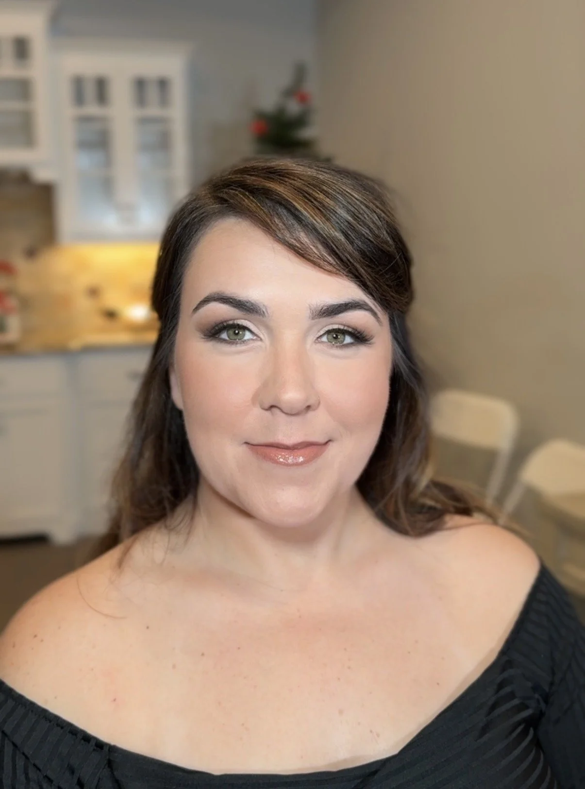 A woman with shoulder-length brown hair and light makeup, wearing an off-the-shoulder black top, smiling in a cozy kitchen with white cabinets and a small Christmas tree in the background.