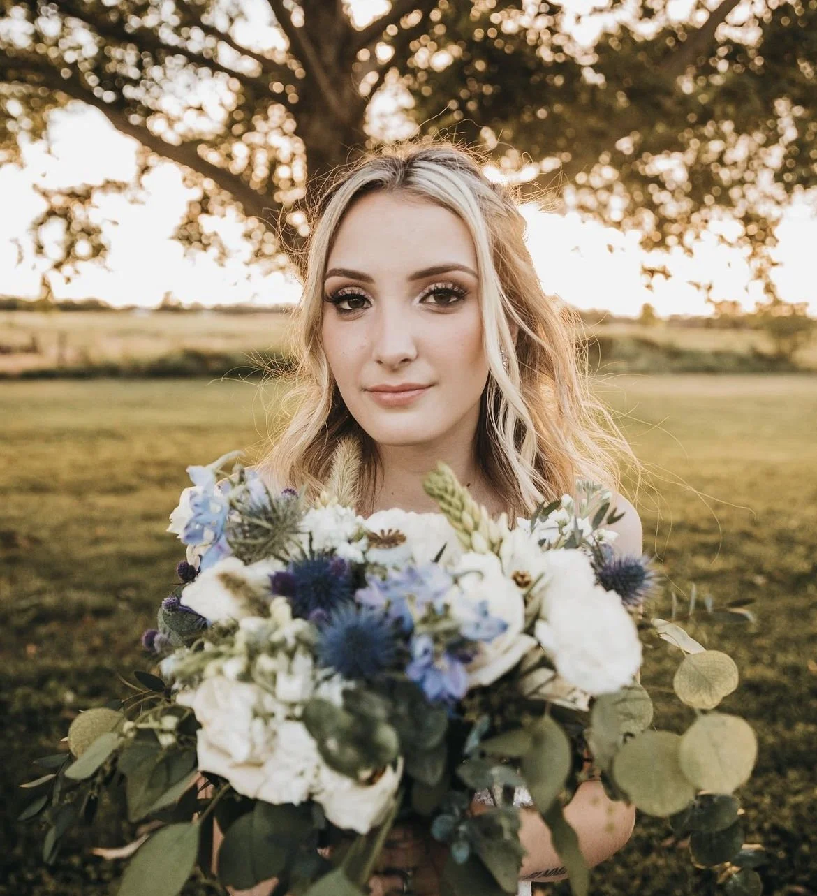A young woman with long blonde hair holding a bouquet of white and blue flowers outdoors at sunset, standing in front of a large tree.