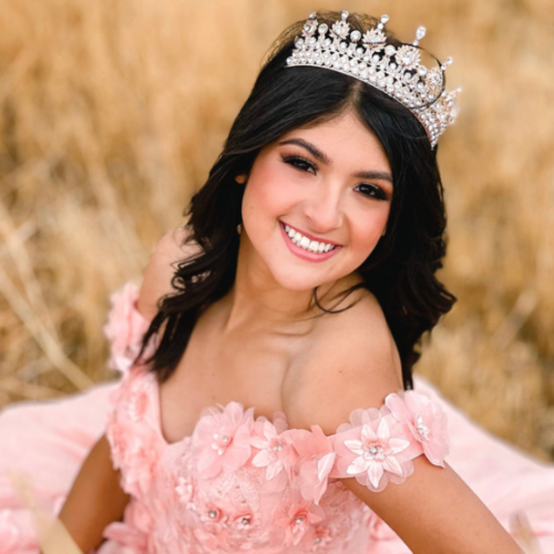 A woman wearing a tiara and a pink off-shoulder dress with floral embellishments smiling outdoors with a blurred background.