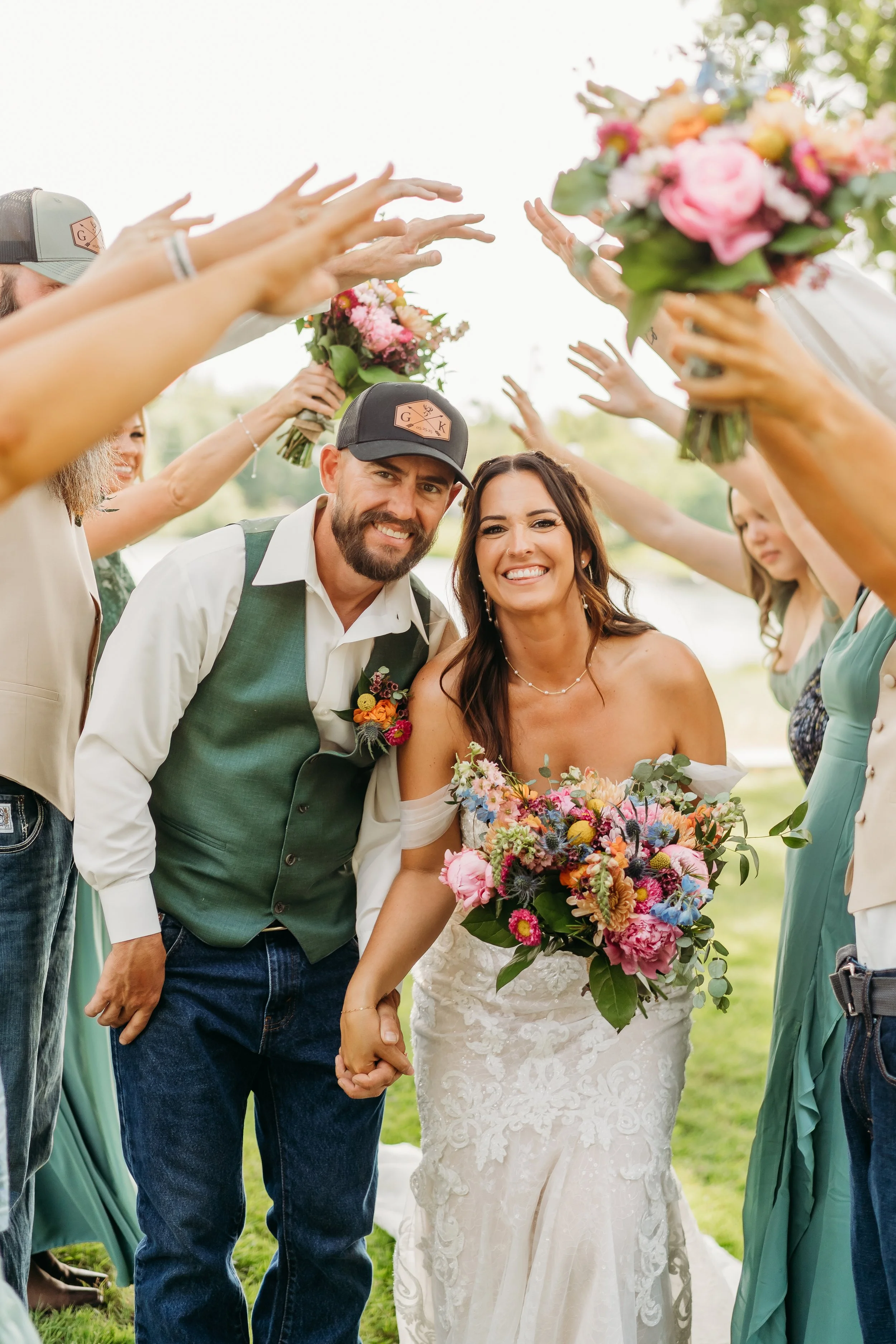 A newlywed couple holding hands, smiling, and holding a bouquet, surrounded by friends with outstretched arms holding smaller bouquets during an outdoor wedding celebration.