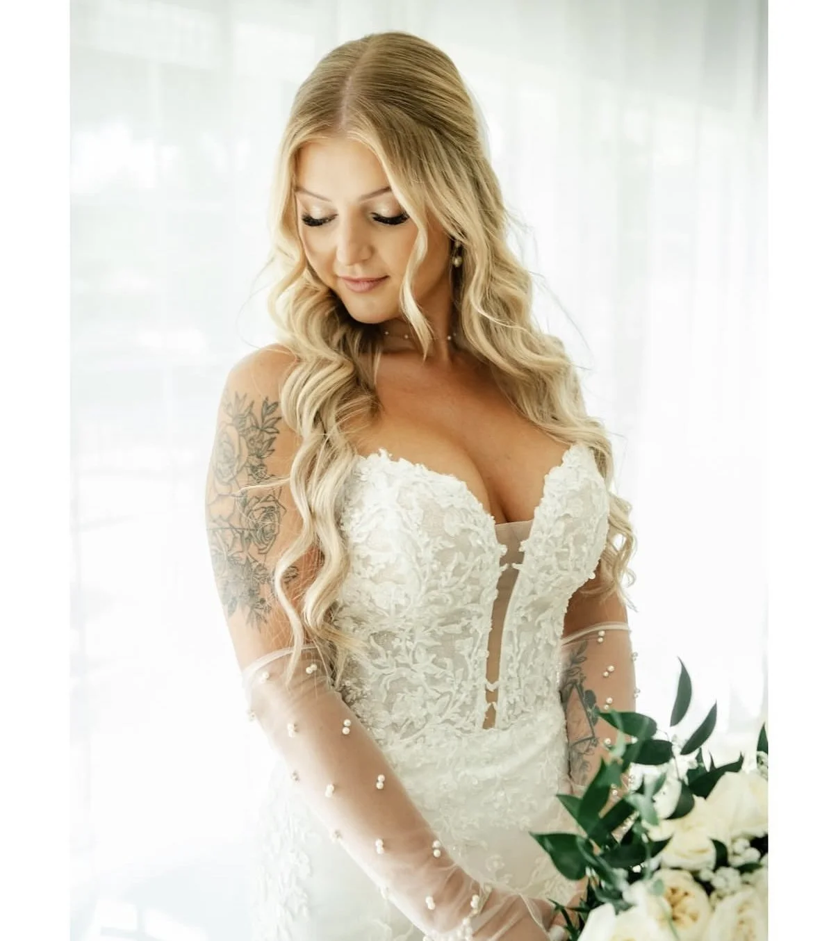 A bride with long blonde hair wearing a white lace wedding dress with pearl details, holding a bouquet of white flowers and greenery.