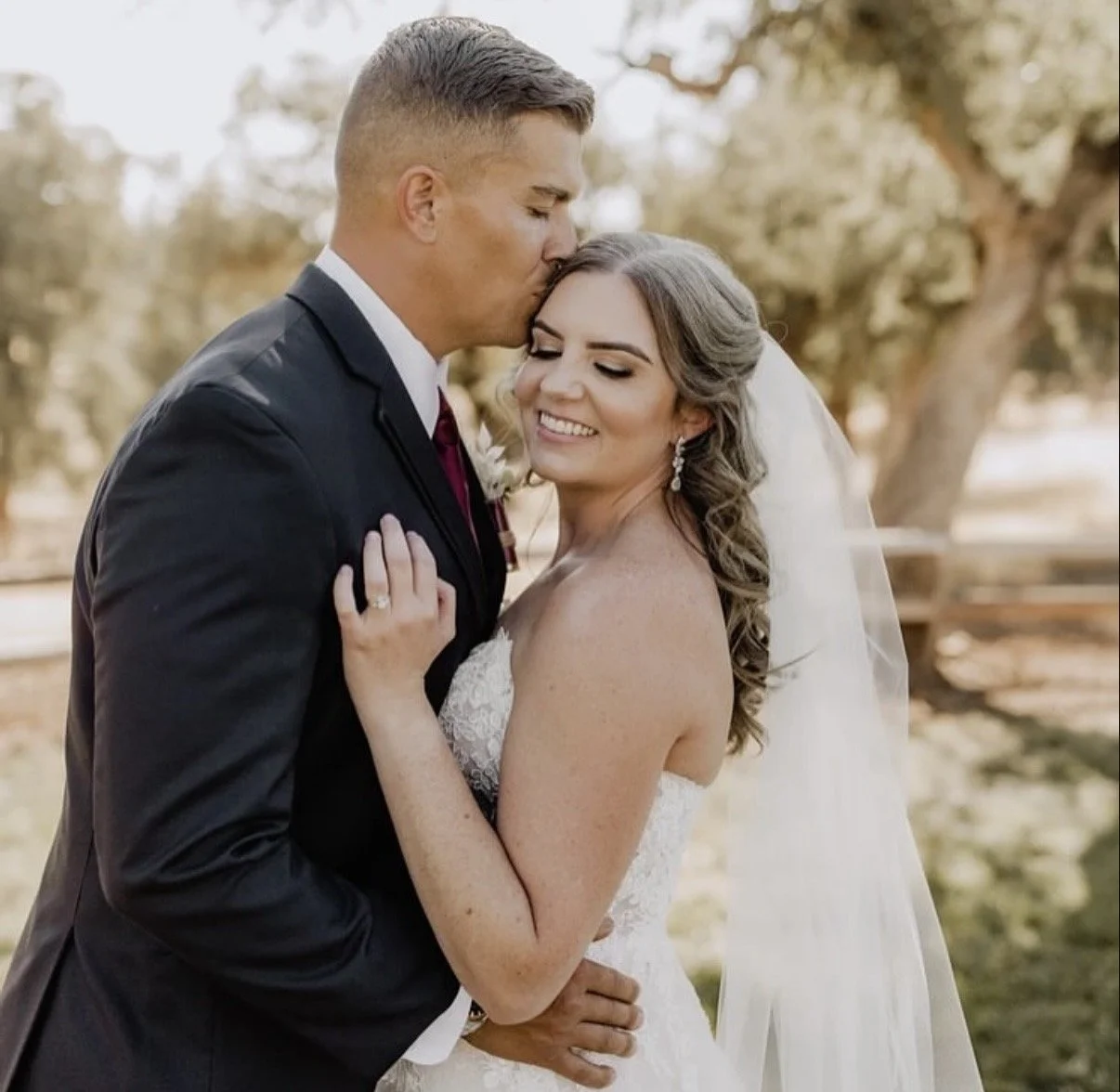 A groom kissing a bride on the forehead while holding her, outdoors with trees in the background, during a wedding photo shoot.