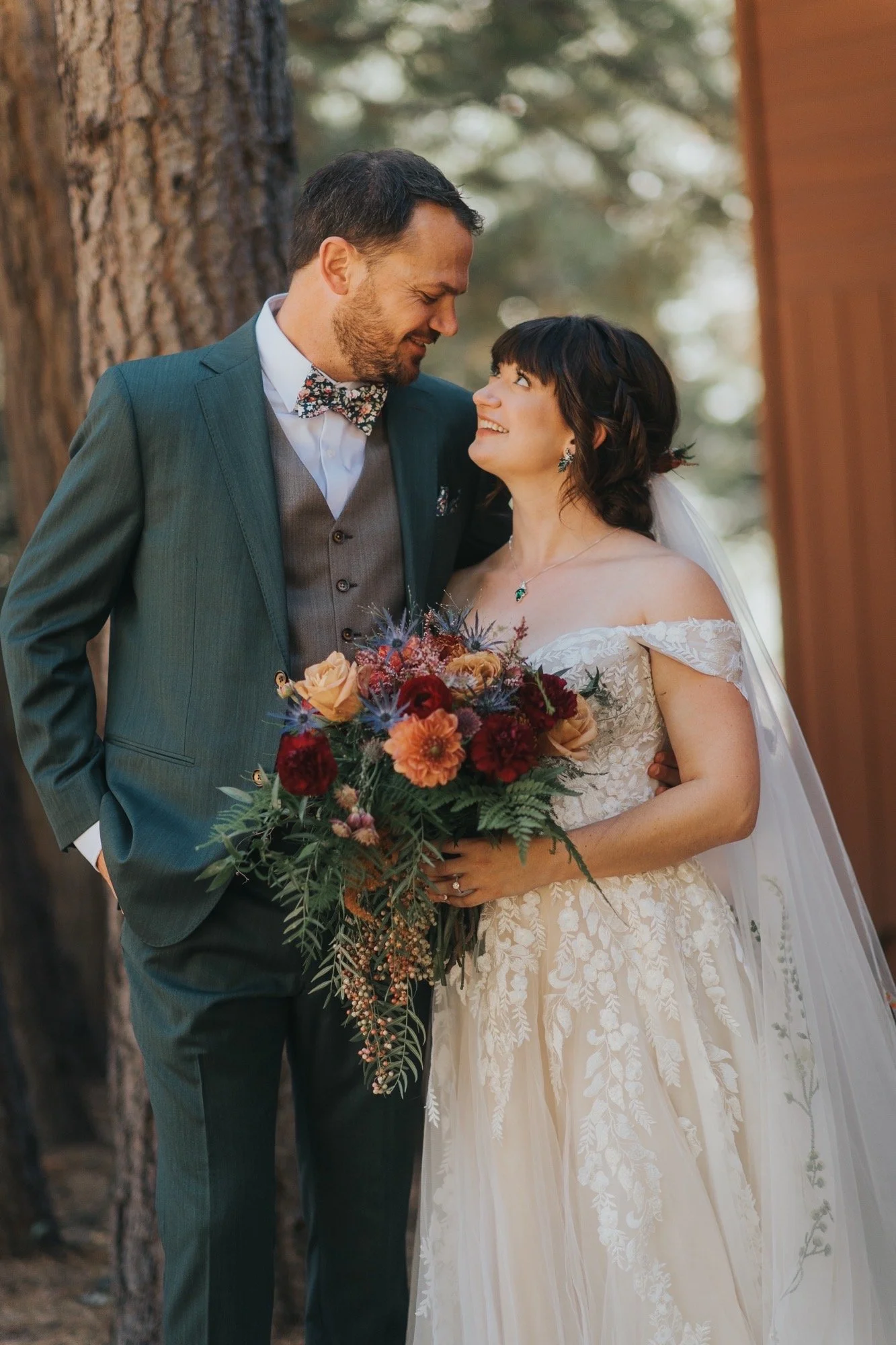 A bride and groom in wedding attire looking at each other outdoors, with the bride holding a bouquet of flowers.