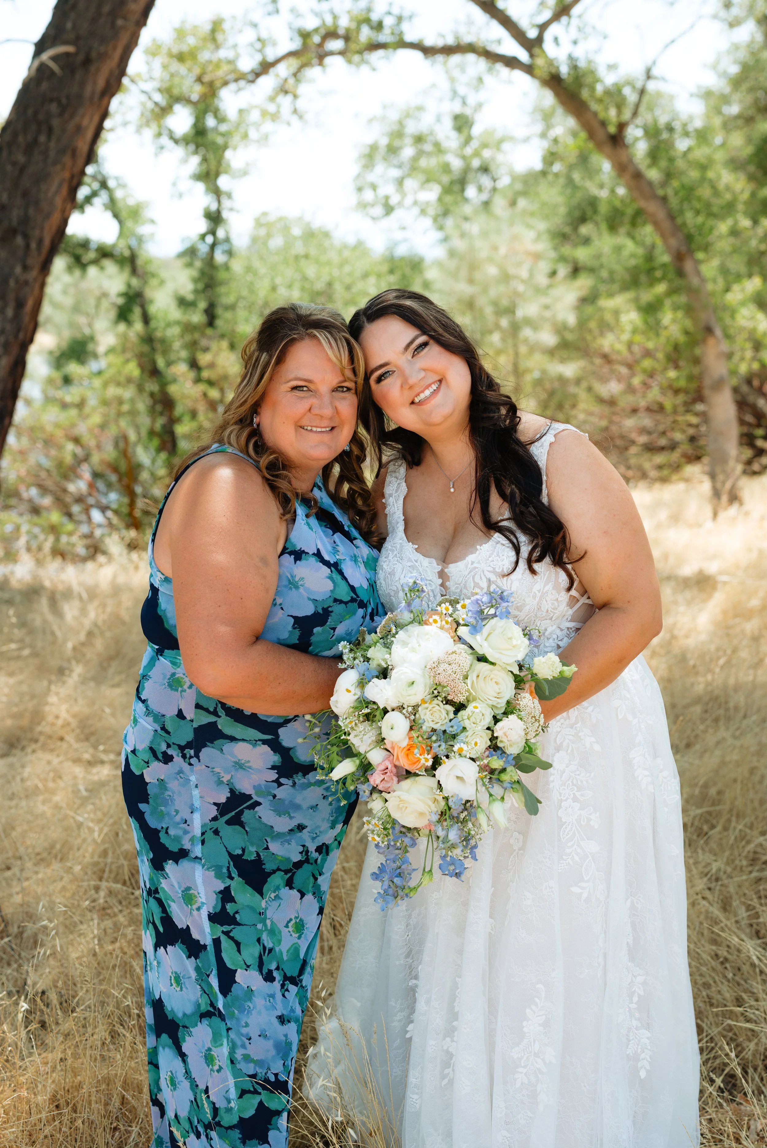 Two women standing outdoors, one in a floral dress and the other in a wedding gown, smiling and holding a bouquet of flowers.