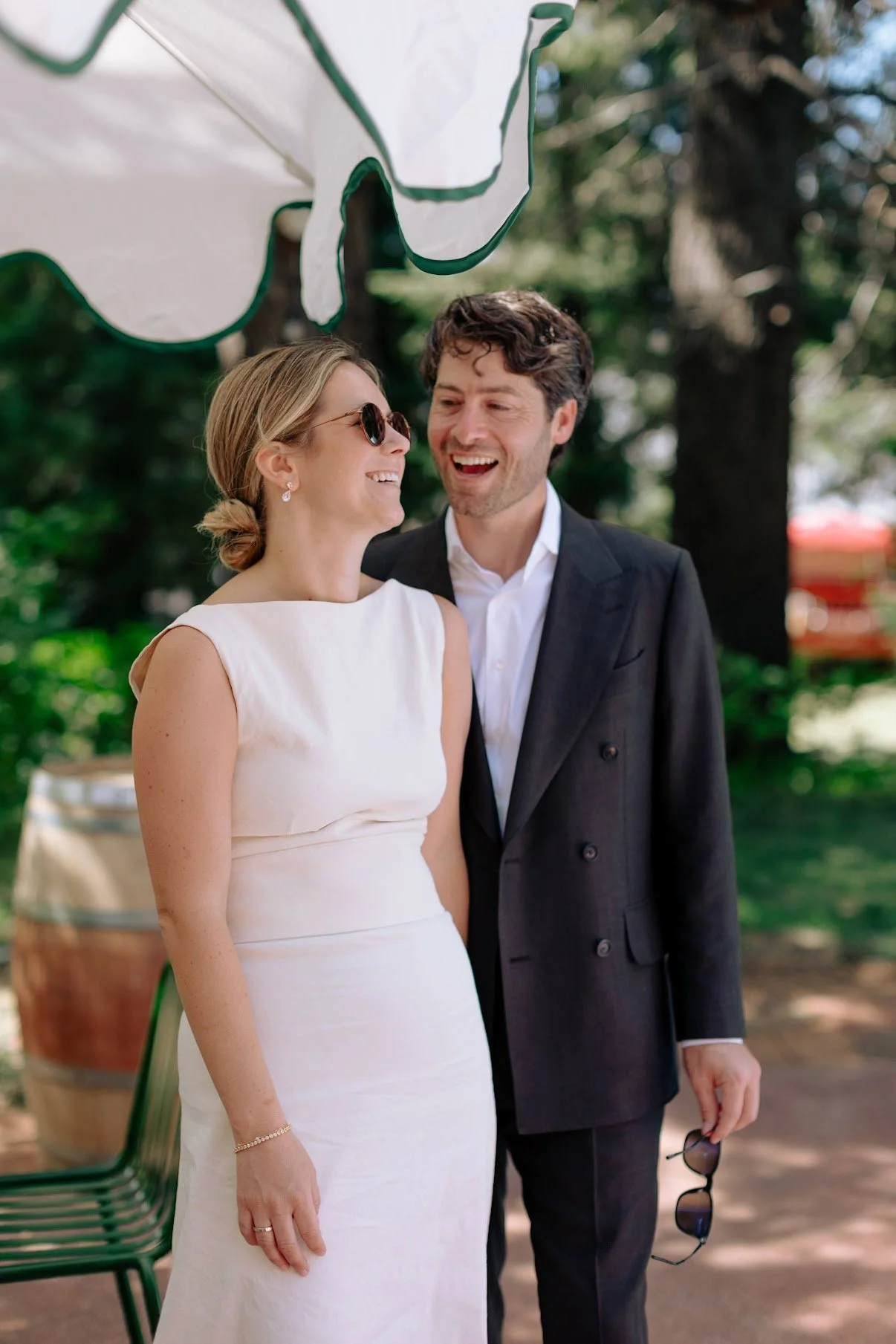A smiling woman in a white dress and sunglasses is standing next to a man in a dark suit, outdoors with trees and a barrel in the background.