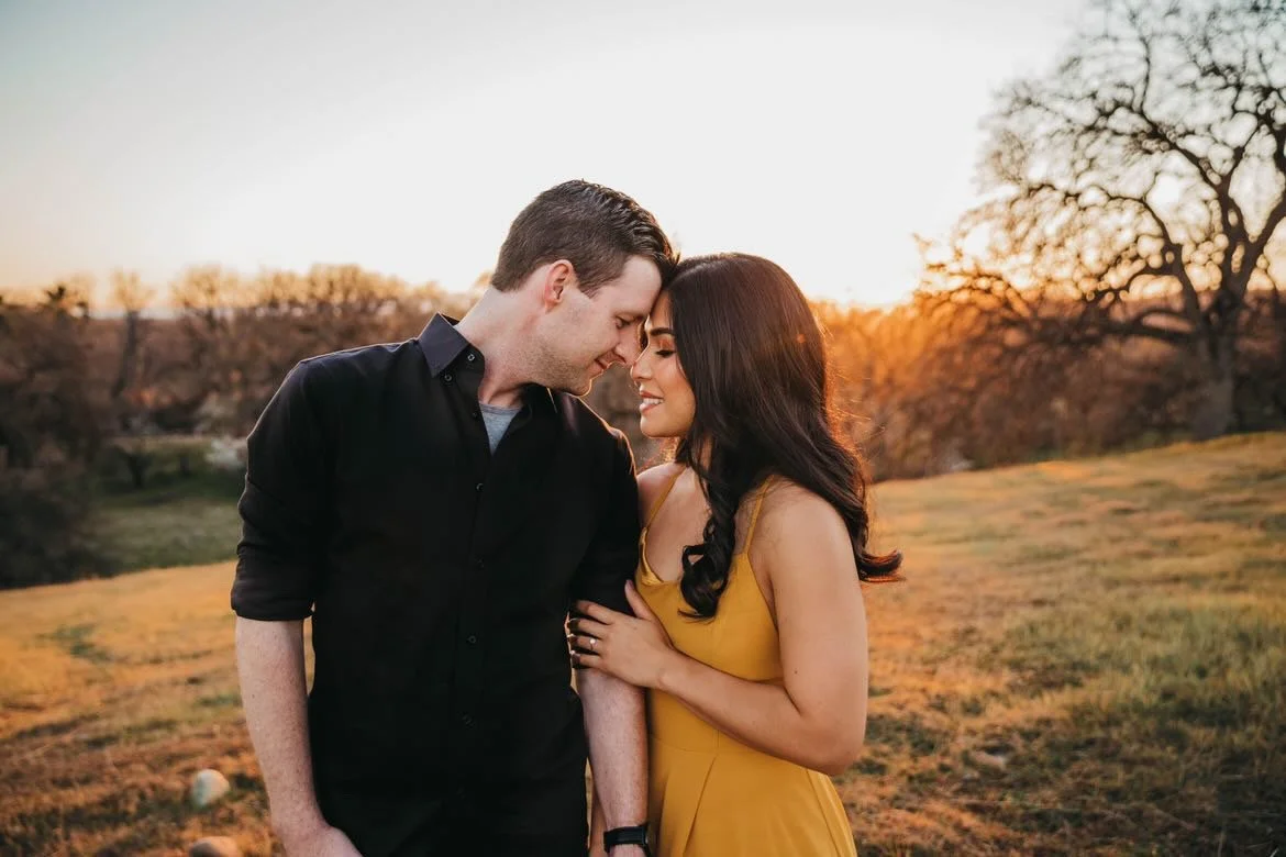 A couple sharing a tender moment outdoors during sunset, with trees and grass in the background.