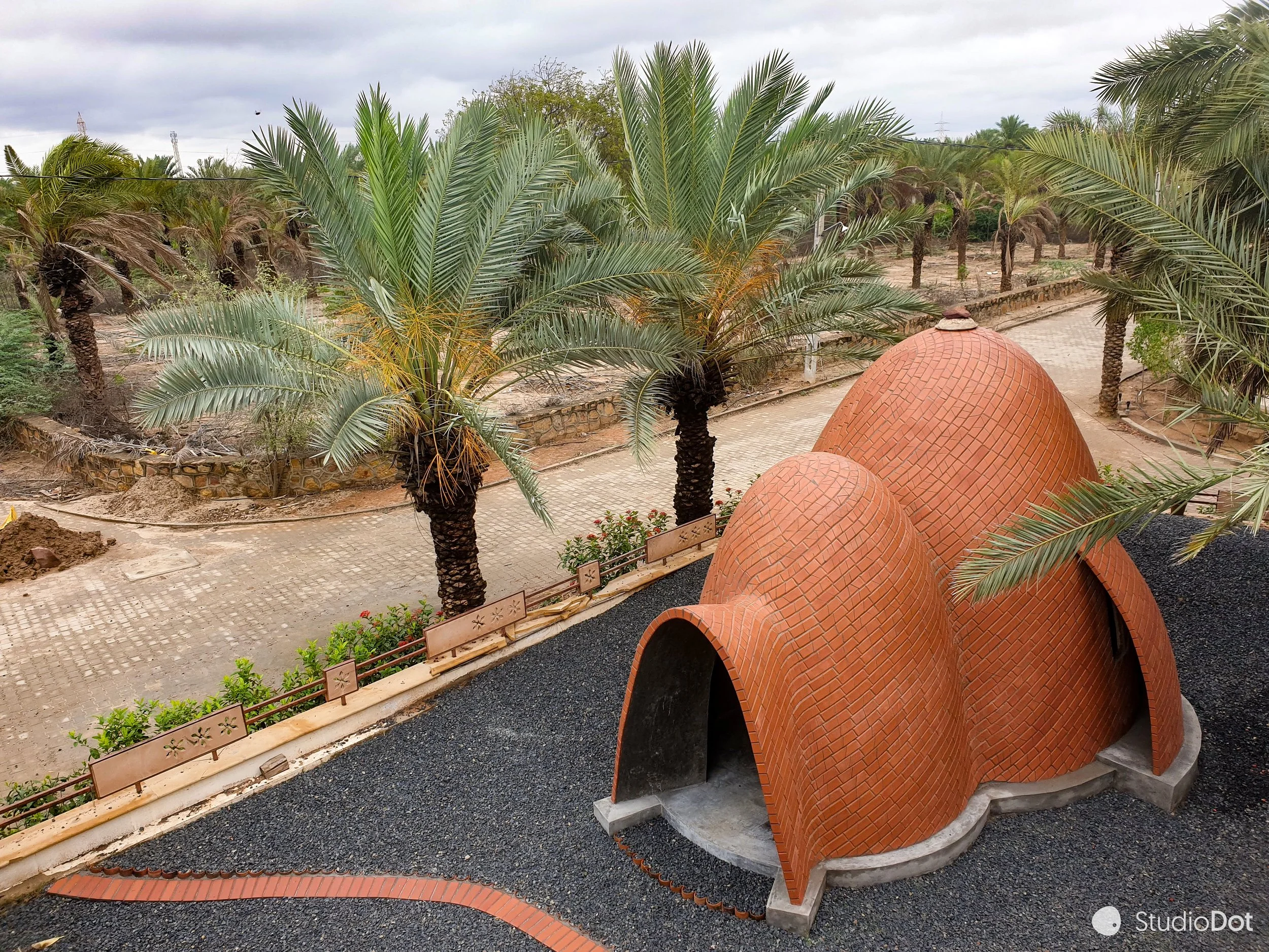 A small, brick, dome-shaped building with three connected domes and arched entrances, set in a landscaped area with palm trees, shrubs, and a paved pathway.